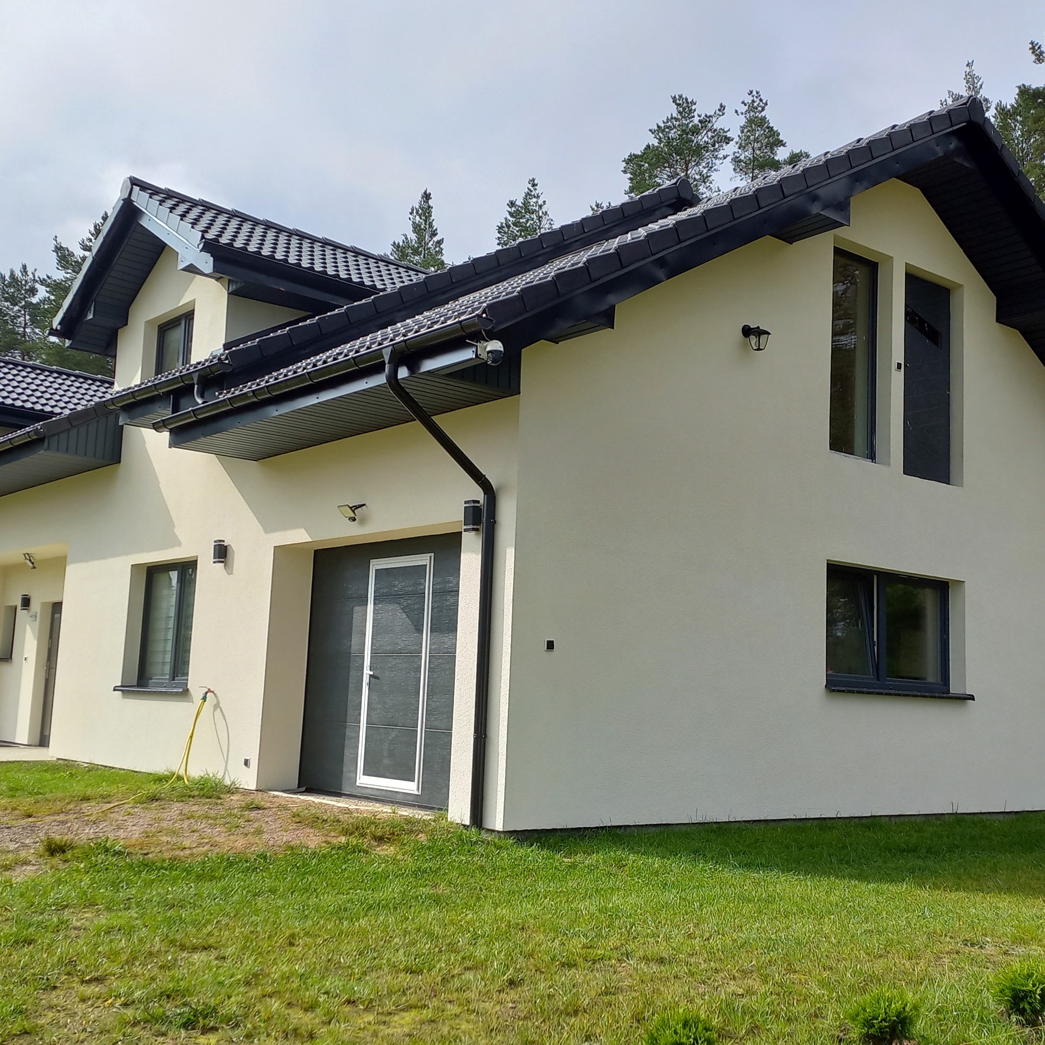 Elewacja domu jednorodzinnego w jasnym odcieniu z ciemną stolarką okienną i dachem. Widoczny fragment trawnika i węża ogrodowego. Ujęcie z dołu, podkreślające bryłę budynku.