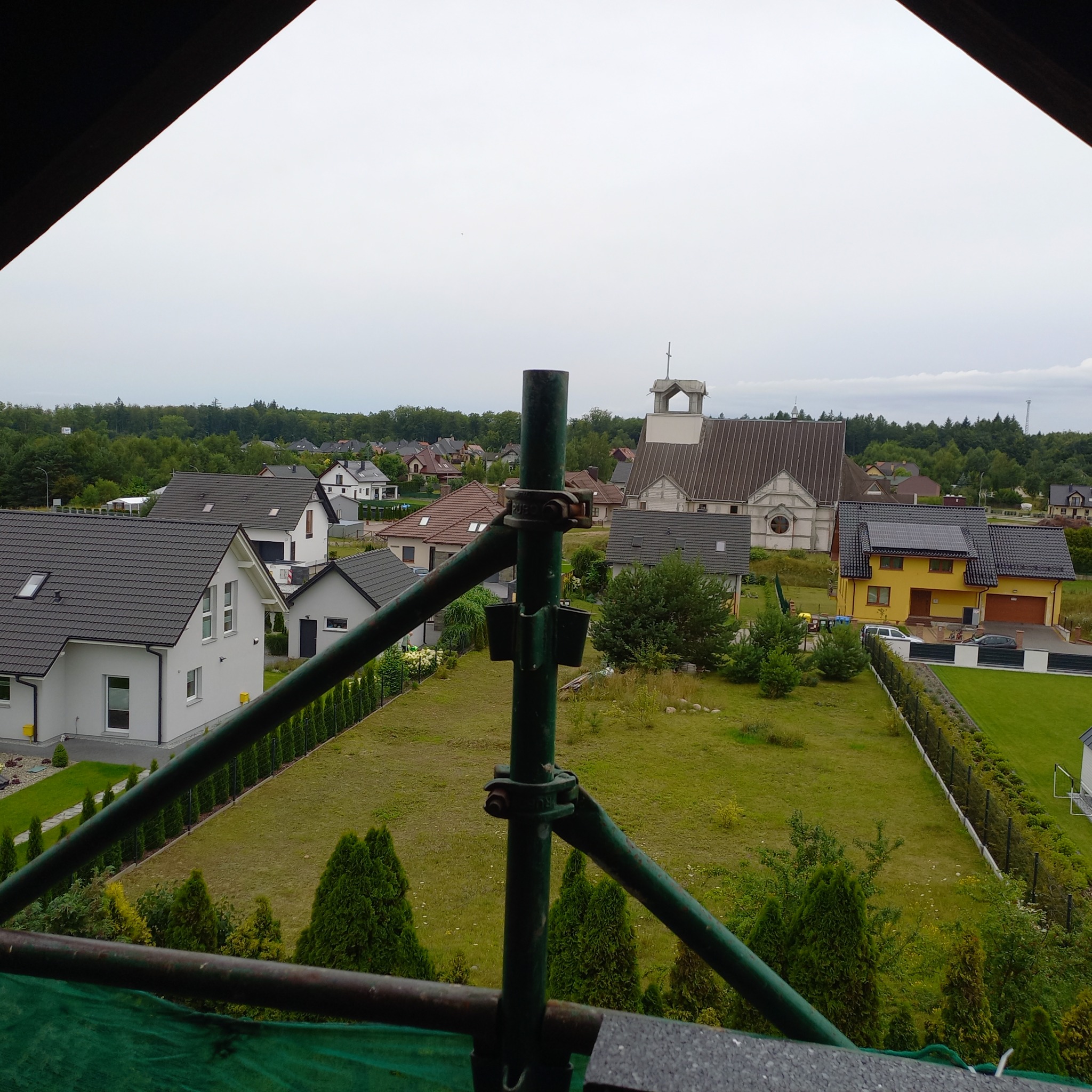 Widok z rusztowania na domy jednorodzinne i kościół w oddali, pochmurne niebo. Elementy rusztowania na pierwszym planie.