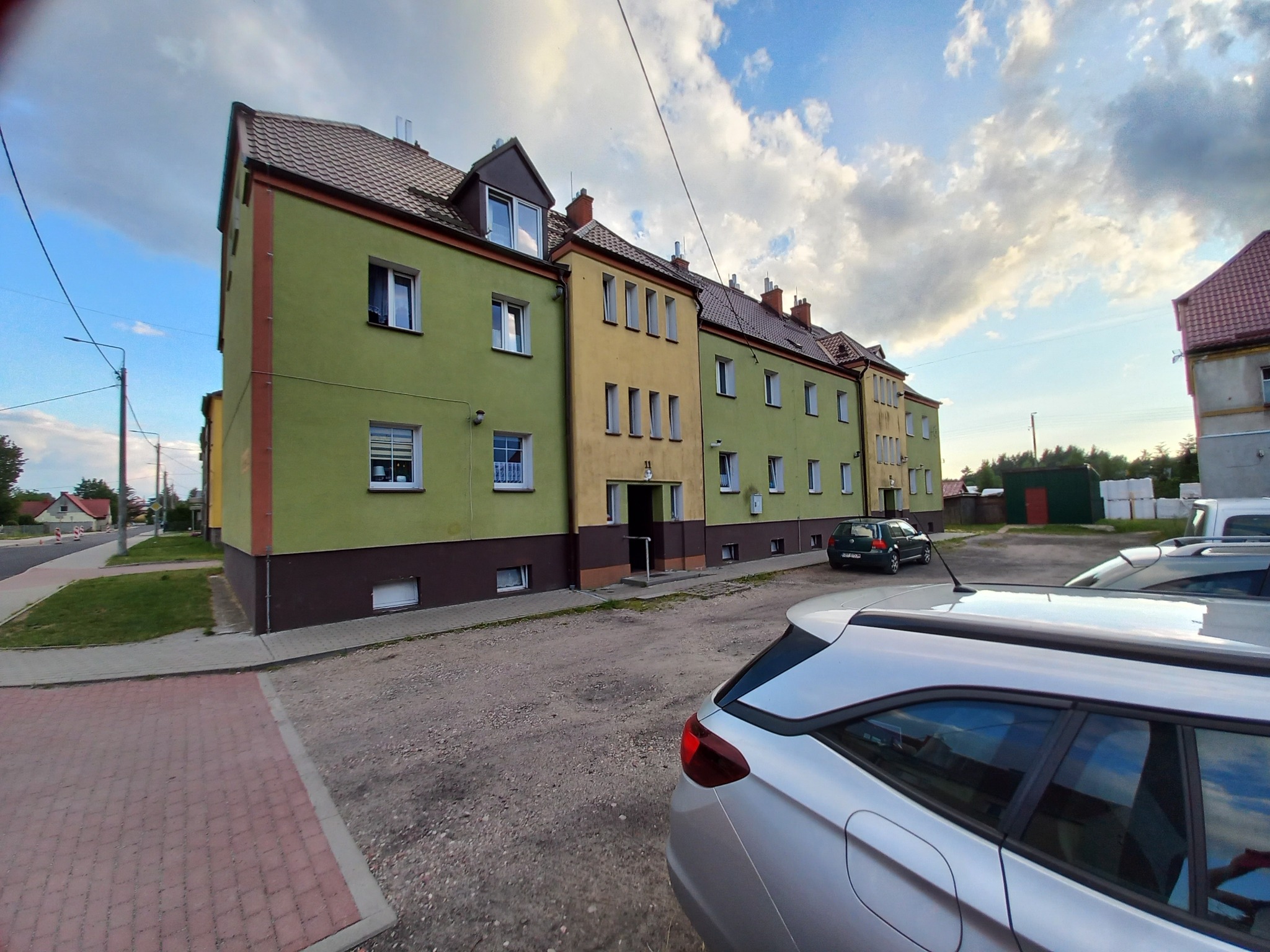 Odnowiony budynek wielorodzinny z zielono-żółtą elewacją i brązowym cokołem, widoczny z perspektywy ulicy, z zaparkowanymi samochodami na pierwszym planie.