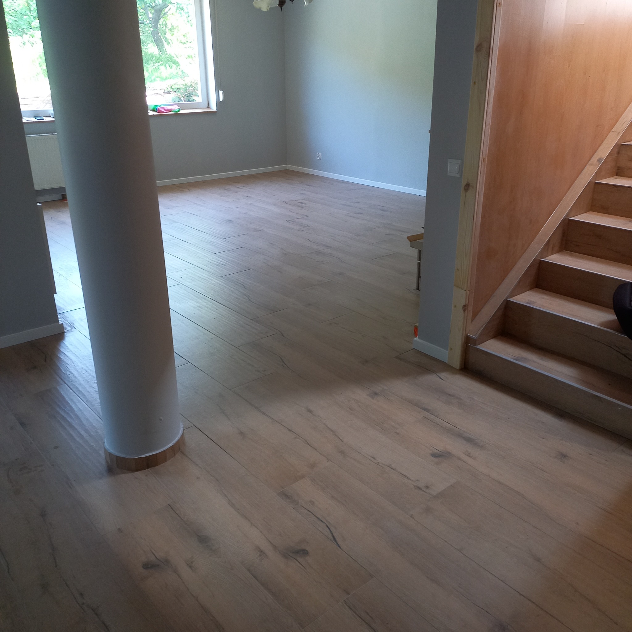 Jasne panele podłogowe ułożone w przestronnym wnętrzu z kolumną i drewnianymi schodami. Świeżo wykończone ściany w odcieniach szarości i widok na zieleń za oknem.