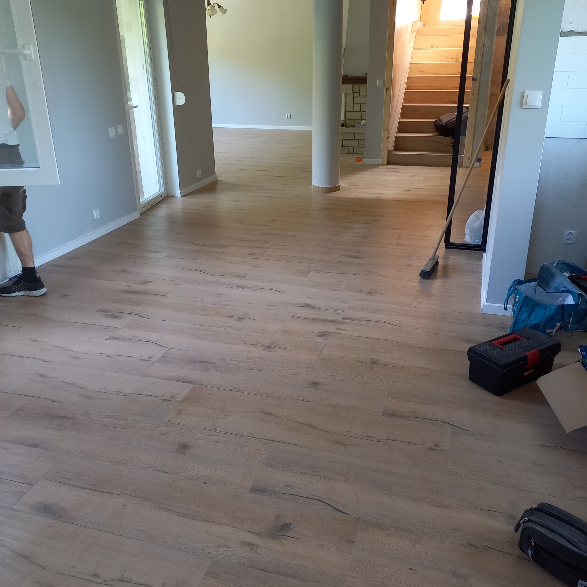 Jasne panele podłogowe z widoczną strukturą drewna w przestronnym wnętrzu domu. W tle schody i kolumna. Częściowo widoczny pracownik i narzędzia.