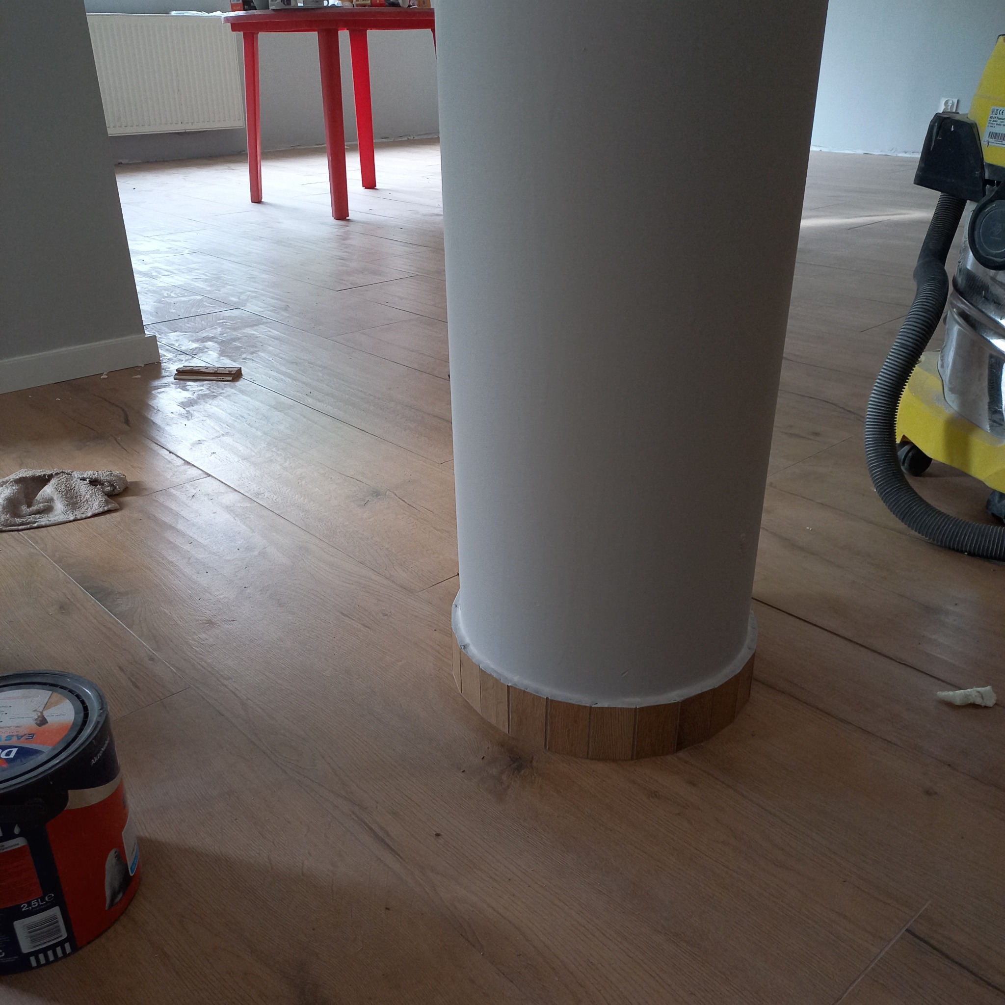Malowanie kolumny w pokoju z drewnianą podłogą. Widoczna puszka farby, odkurzacz i czerwony stolik w tle. Detal wykończenia kolumny z drewnianą podstawą.