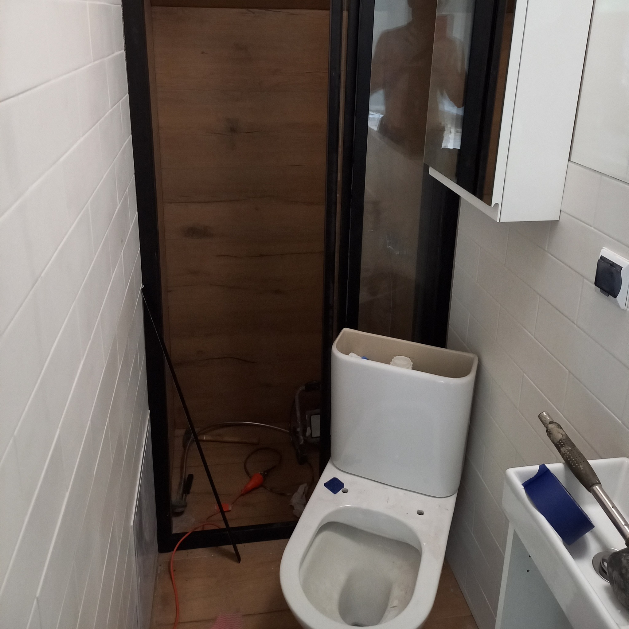 Biała toaleta w trakcie montażu w łazience z białymi, prostokątnymi płytkami. Widoczne narzędzia i instalacja. Ujęcie z bliska, z widocznym fragmentem umywalki.