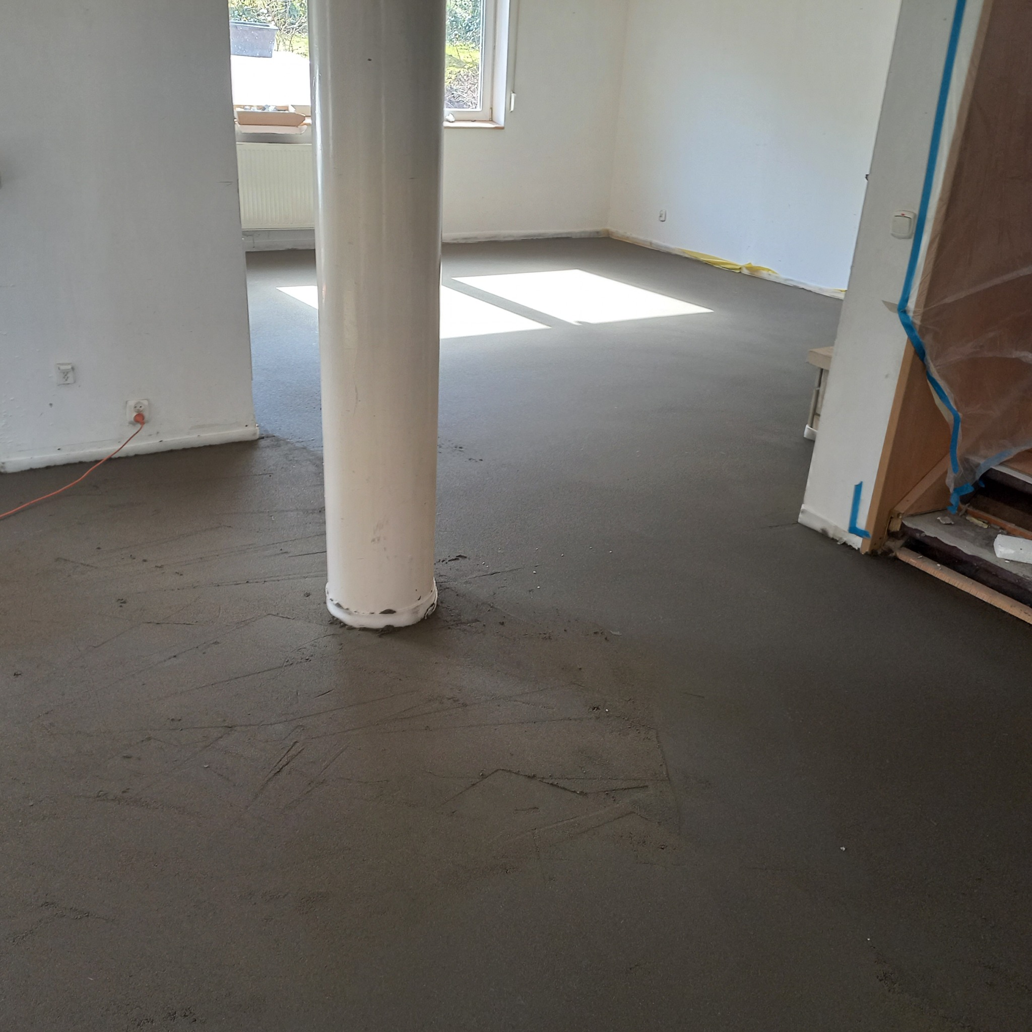 Wnętrze z wylaną, świeżą wylewką podłogową, widoczny filar konstrukcyjny i fragment okna z naturalnym światłem. Przewód elektryczny przy ścianie.
