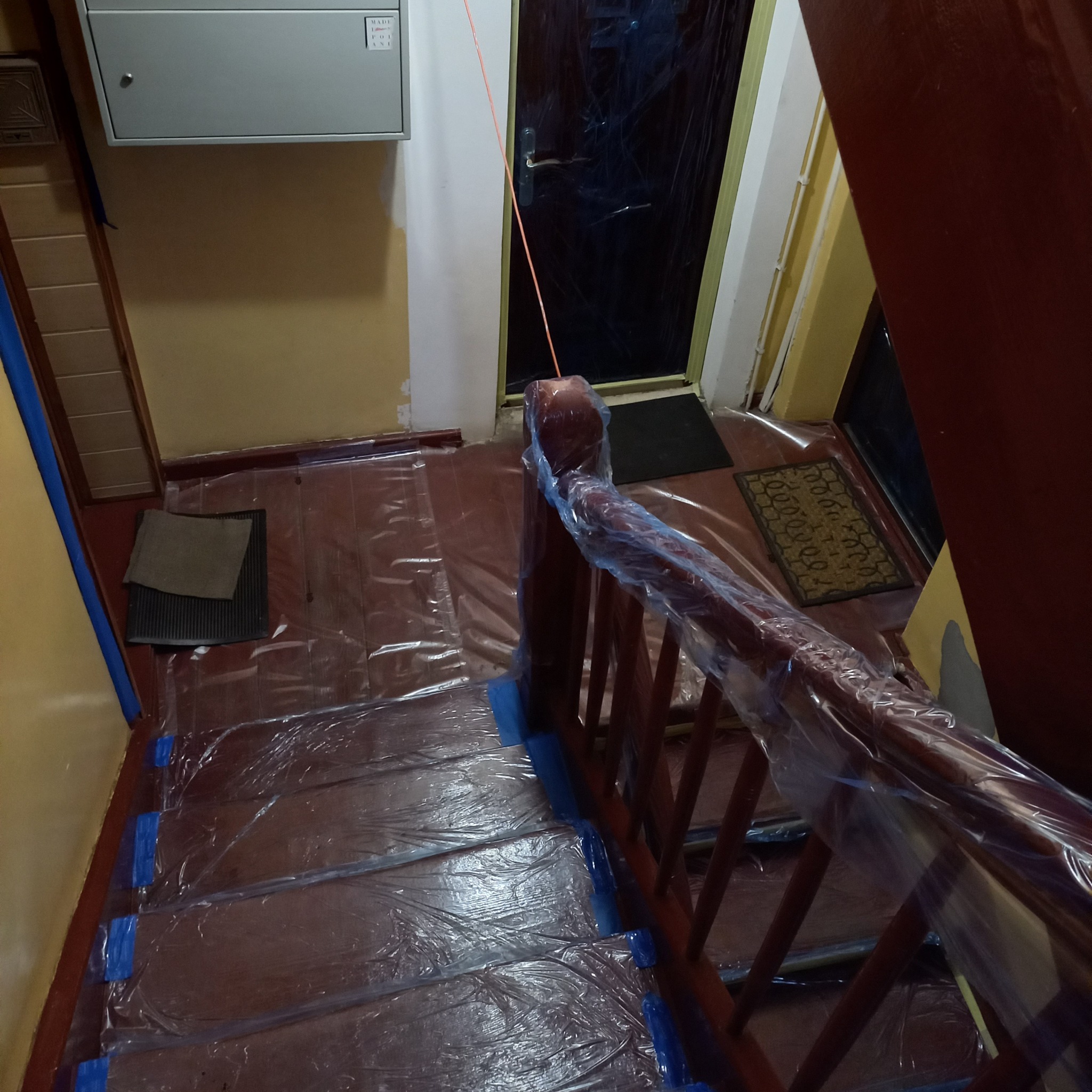 Schody zabezpieczone folią i taśmą malarską przed remontem. Widok z góry na drewniane schody i poręcze przygotowane do prac wykończeniowych wewnątrz budynku.