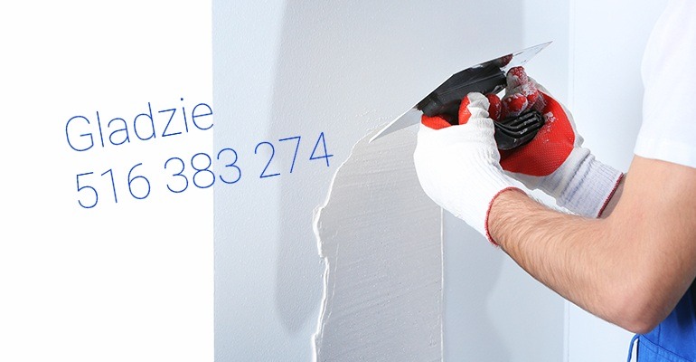 Nakładanie gładzi na ścianę za pomocą szpachelki. Widoczne ręce w rękawicach i fragment ściany z nałożoną gładzią. Na ścianie numer telefonu do wykonawcy.