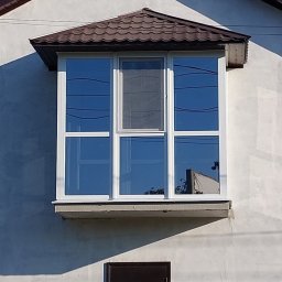 Oleksandr Kozachenko - Wystający balkon z nowymi, białymi oknami z brązowym daszkiem, widoczne odbicie drzewa w szybach.