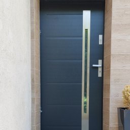 Oleksandr Kozachenko - Ciemnoszare drzwi wejściowe z pionowym, metalowym panelem i szybą, zamontowane w ościeżnicy wykończonej kafelkami imitującymi drewno.