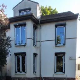 Dwupiętrowy dom z nowymi oknami i roletami zewnętrznymi, w trakcie wykończenia elewacji, widoczne gruz i materiały budowlane.