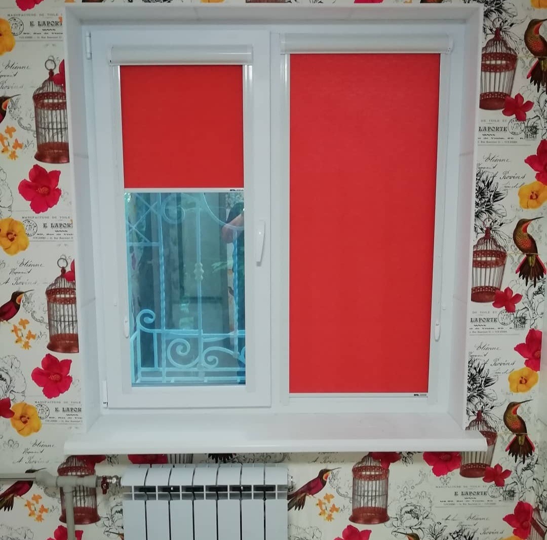 Białe okno PCV z dwoma skrzydłami, jedno z częściowo opuszczoną czerwoną roletą, drugie z widoczną kratą w oknie, na tle kwiecistej tapety z motywem klatek dla ptaków.