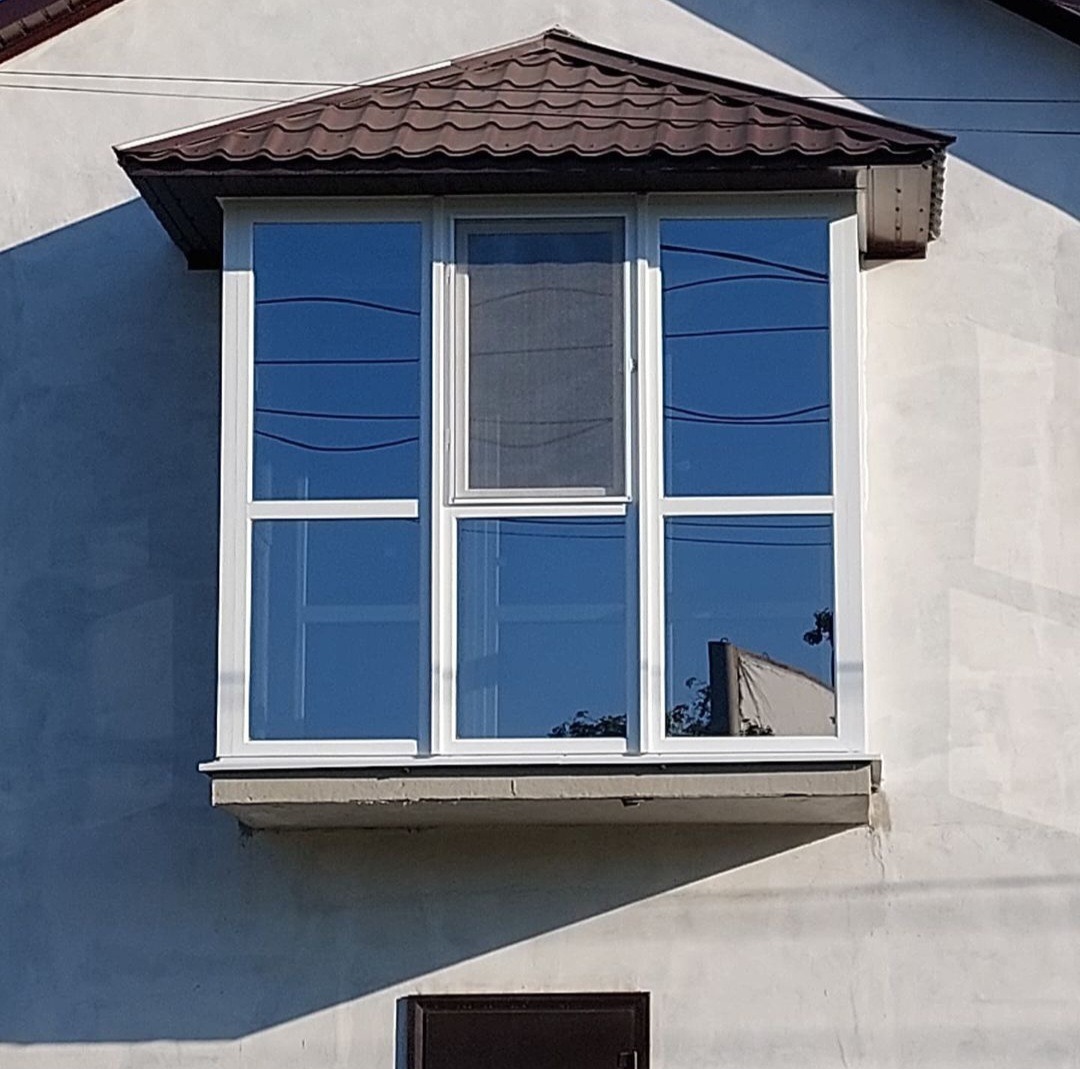 Wystający balkon z nowymi, białymi oknami z brązowym daszkiem, widoczne odbicie drzewa w szybach.