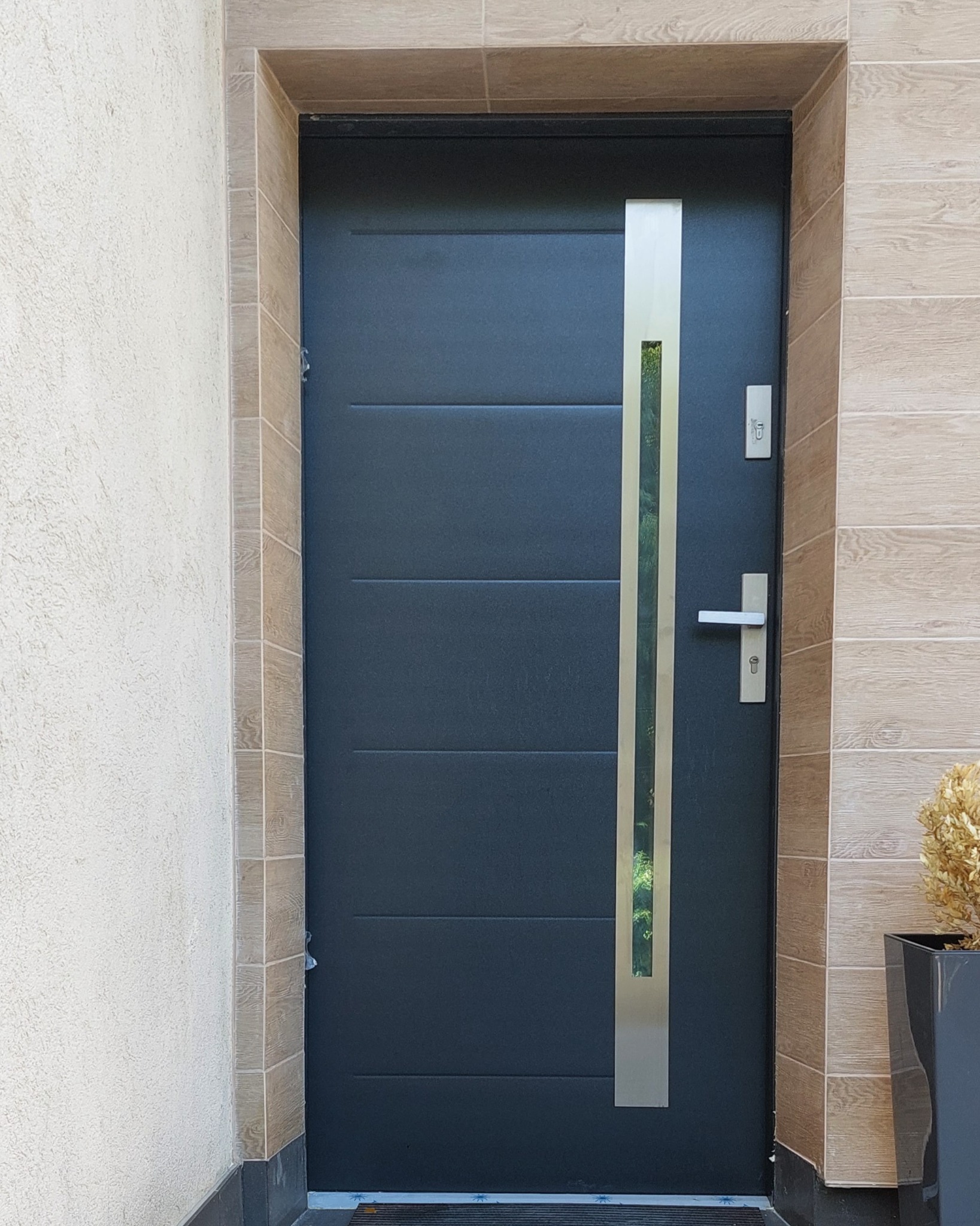 Ciemnoszare drzwi wejściowe z pionowym, metalowym panelem i szybą, zamontowane w ościeżnicy wykończonej kafelkami imitującymi drewno.