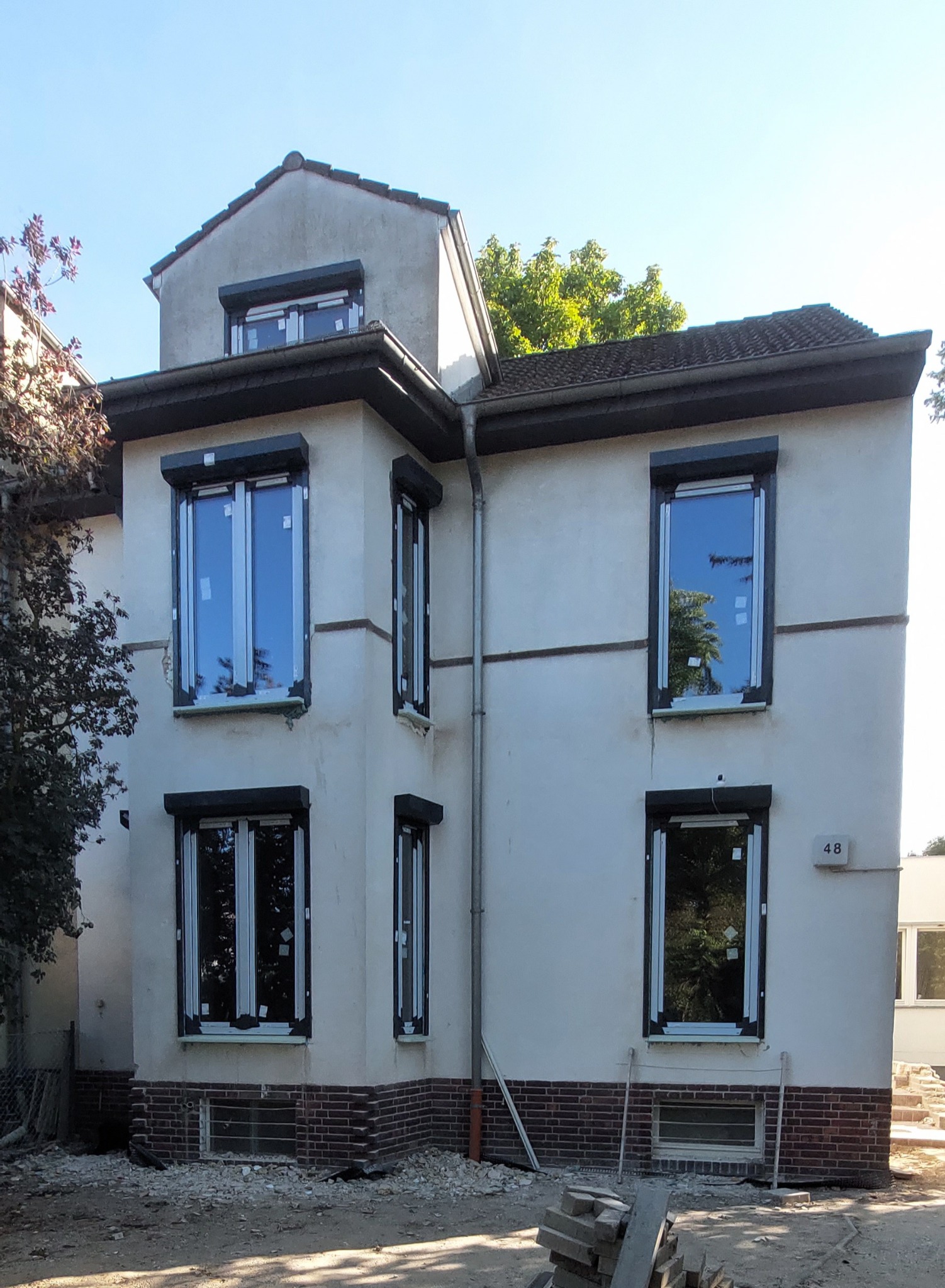 Dwupiętrowy dom z nowymi oknami i roletami zewnętrznymi, w trakcie wykończenia elewacji, widoczne gruz i materiały budowlane.