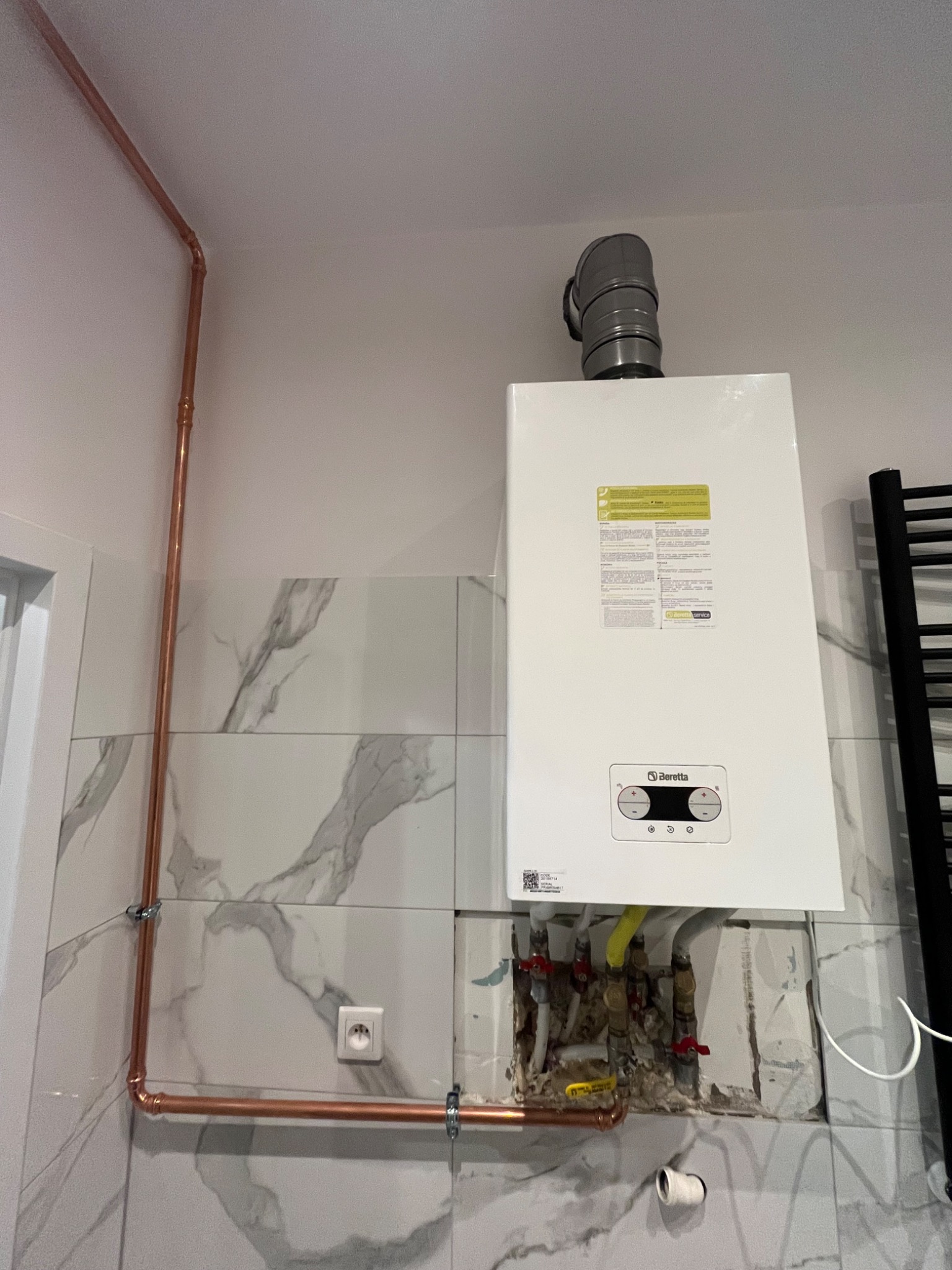 Biały piec gazowy marki Beretta zamontowany na ścianie w łazience z marmurowymi płytkami, widoczne miedziane rury instalacji grzewczej i fragment niedokończonej zabudowy pod piecem.