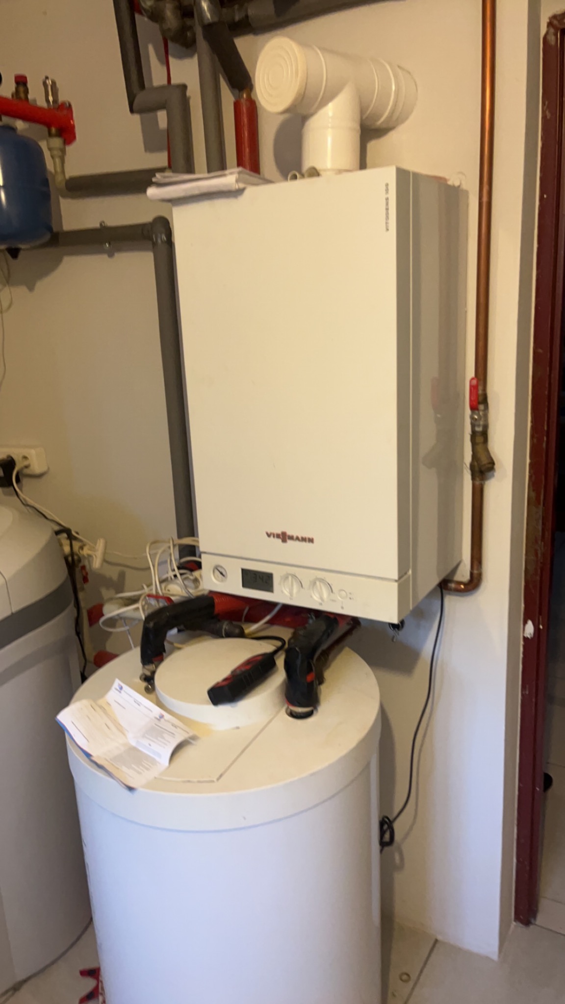 Kocioł gazowy Viessmann zamontowany na białym zbiorniku, widoczne rury doprowadzające, narzędzia na zbiorniku i dokumentacja techniczna obok, na ścianie szare rury instalacyjne.