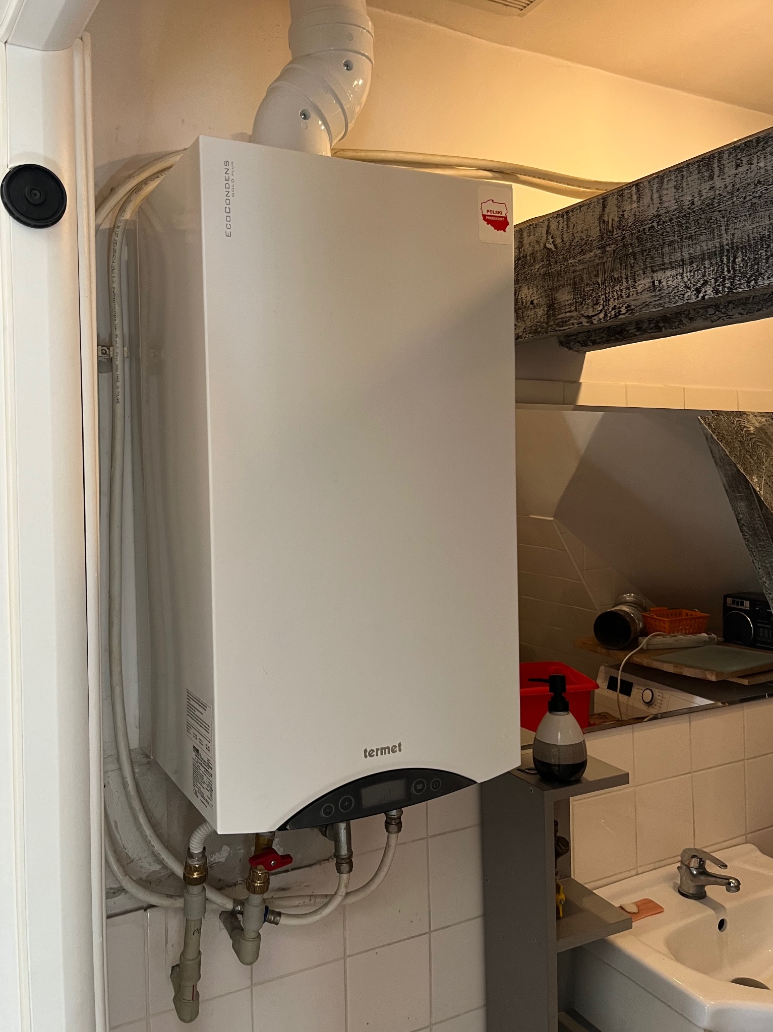 Biały, wiszący piec gazowy marki Termet z podłączonymi rurami i odprowadzeniem spalin, zamontowany na kafelkowanej ścianie w pomieszczeniu z umywalką i drewnianą belką stropową.