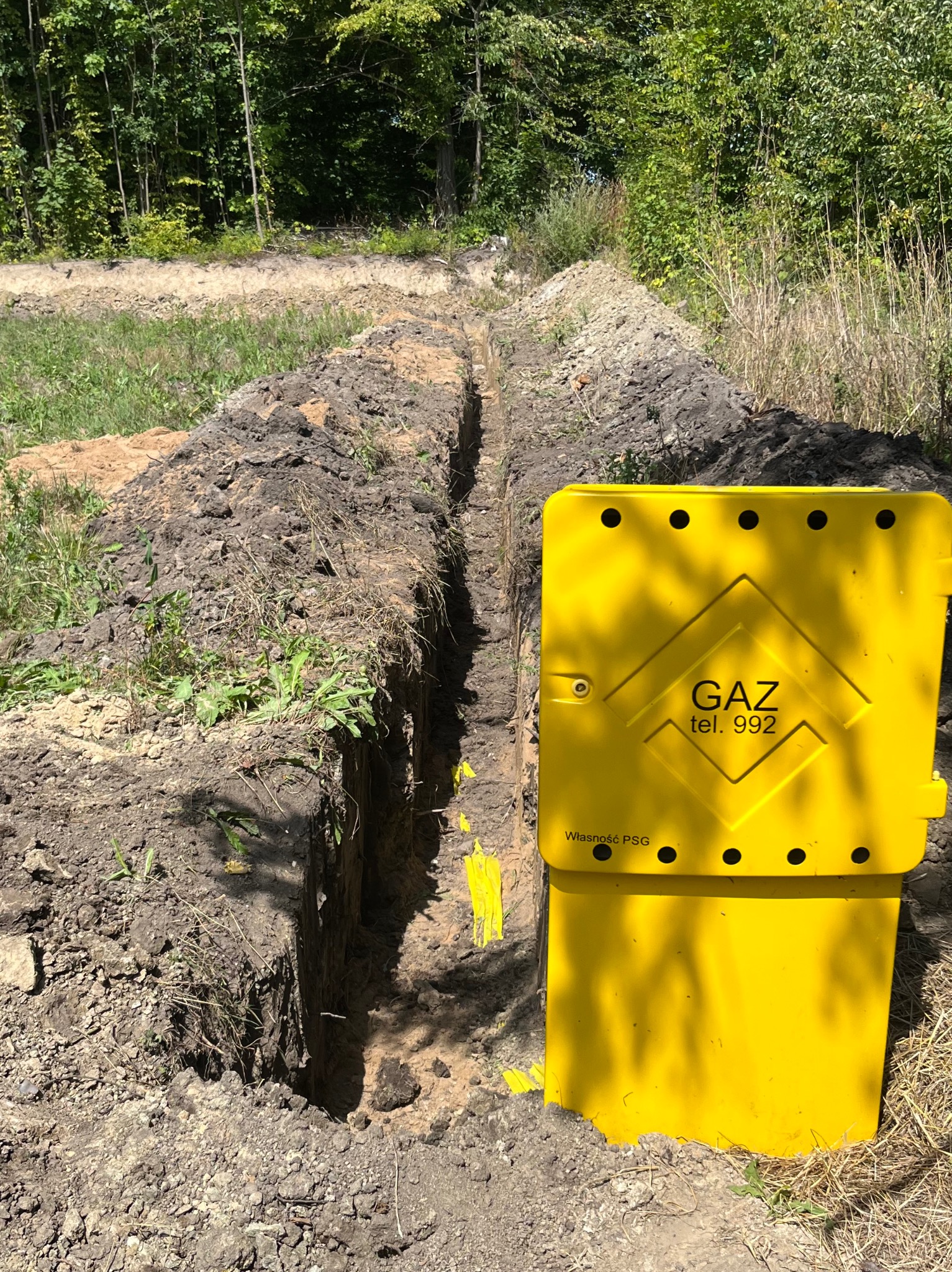 Żółta skrzynka gazowa z napisem 'GAZ tel. 992' stojąca przy wykopanym rowie w ziemi, prawdopodobnie podczas układania instalacji gazowej.