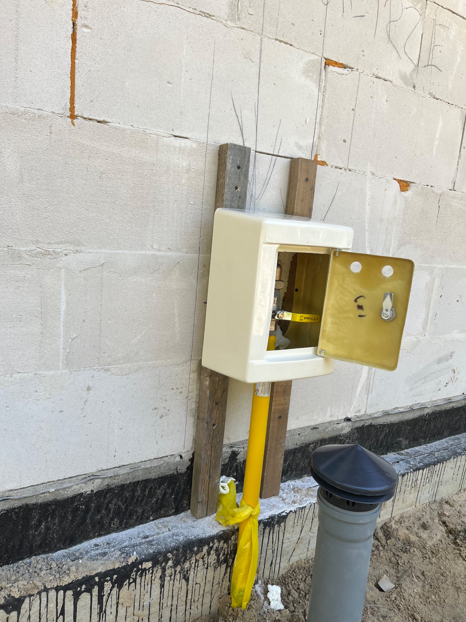 Otwarta skrzynka gazowa z żółtą rurą gazową i zaworem, zamontowana na drewnianych łatach przy szarej ścianie z betonu komórkowego, widoczny komin wentylacyjny i folia w płynie