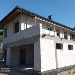 Surowy stan domu z białych bloczków, widoczne rusztowania, czarny dach i orynnowanie, stan surowy otwarty, w tle drzewa i błękitne niebo.