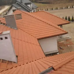 Ceglasty dach z kominem w trakcie budowy, widok z góry. Częściowo położona dachówka, widoczne elementy konstrukcji dachu. W tle dom i teren budowy.