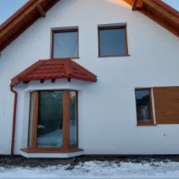 Biały dom jednorodzinny z drewnianą stolarką okienną i czerwoną dachówką, częściowo ocieplony brązowymi panelami elewacyjnymi, zimowy krajobraz.