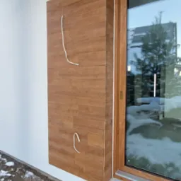 Fragment ściany zewnętrznej domu z drewnopodobną okładziną elewacyjną, widoczne wystające przewody i okno z drewnianą ramą, w tle ogród.