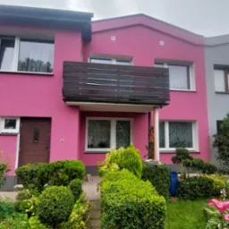 Różowa fasada domu z ciemnym balkonem i zadbanym ogrodem. Widok na dwukondygnacyjny budynek z ciemnymi oknami i brązowymi drzwiami wejściowymi. Pochmurne niebo w tle.