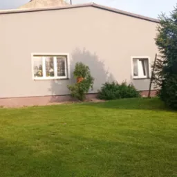 Szary budynek z dwoma oknami, otoczony trawnikiem i zielenią, z widoczną strukturą tynku na elewacji.