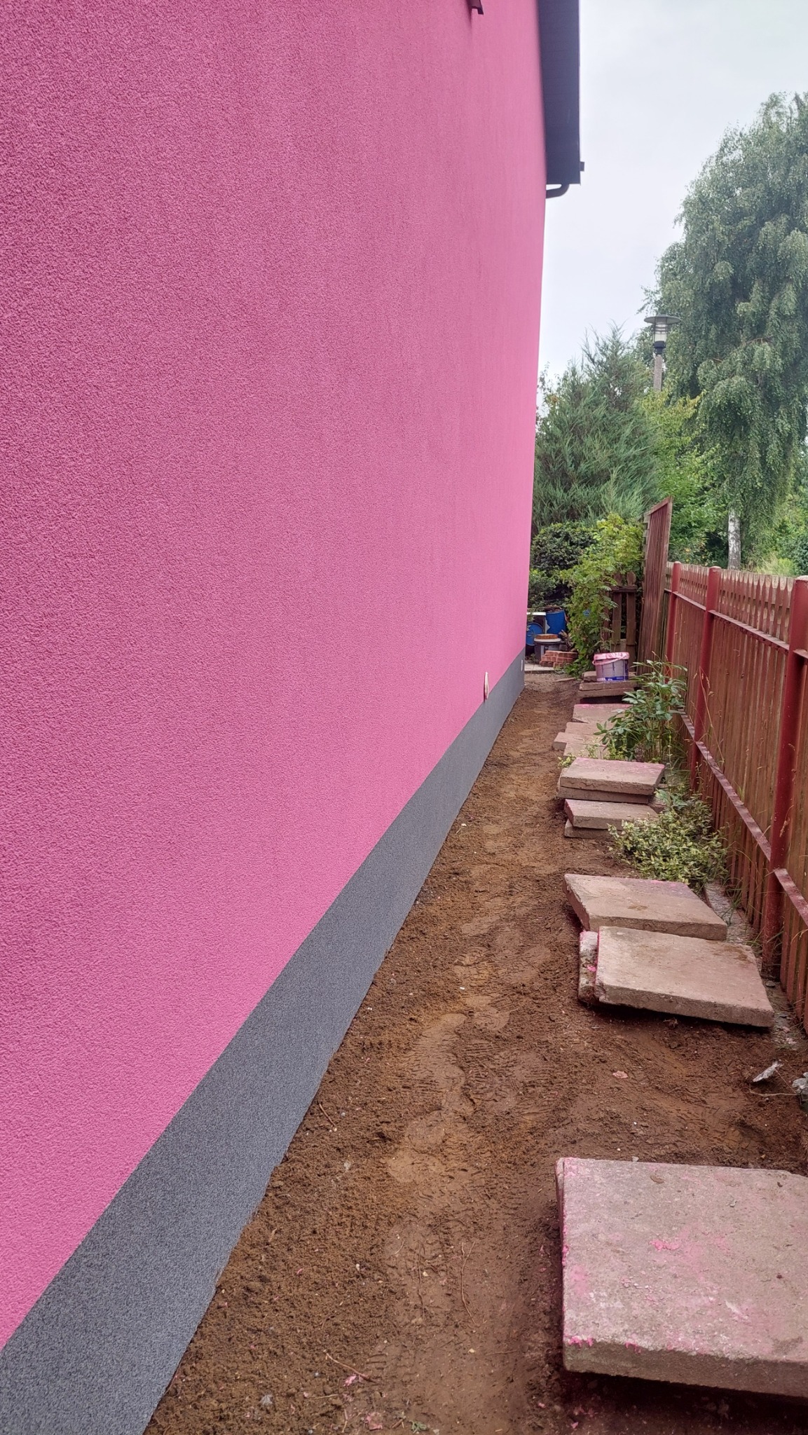 Różowa elewacja domu z szarą podmurówką, widok z boku na ścieżkę z płyt betonowych wzdłuż ściany i drewniane ogrodzenie w tle.