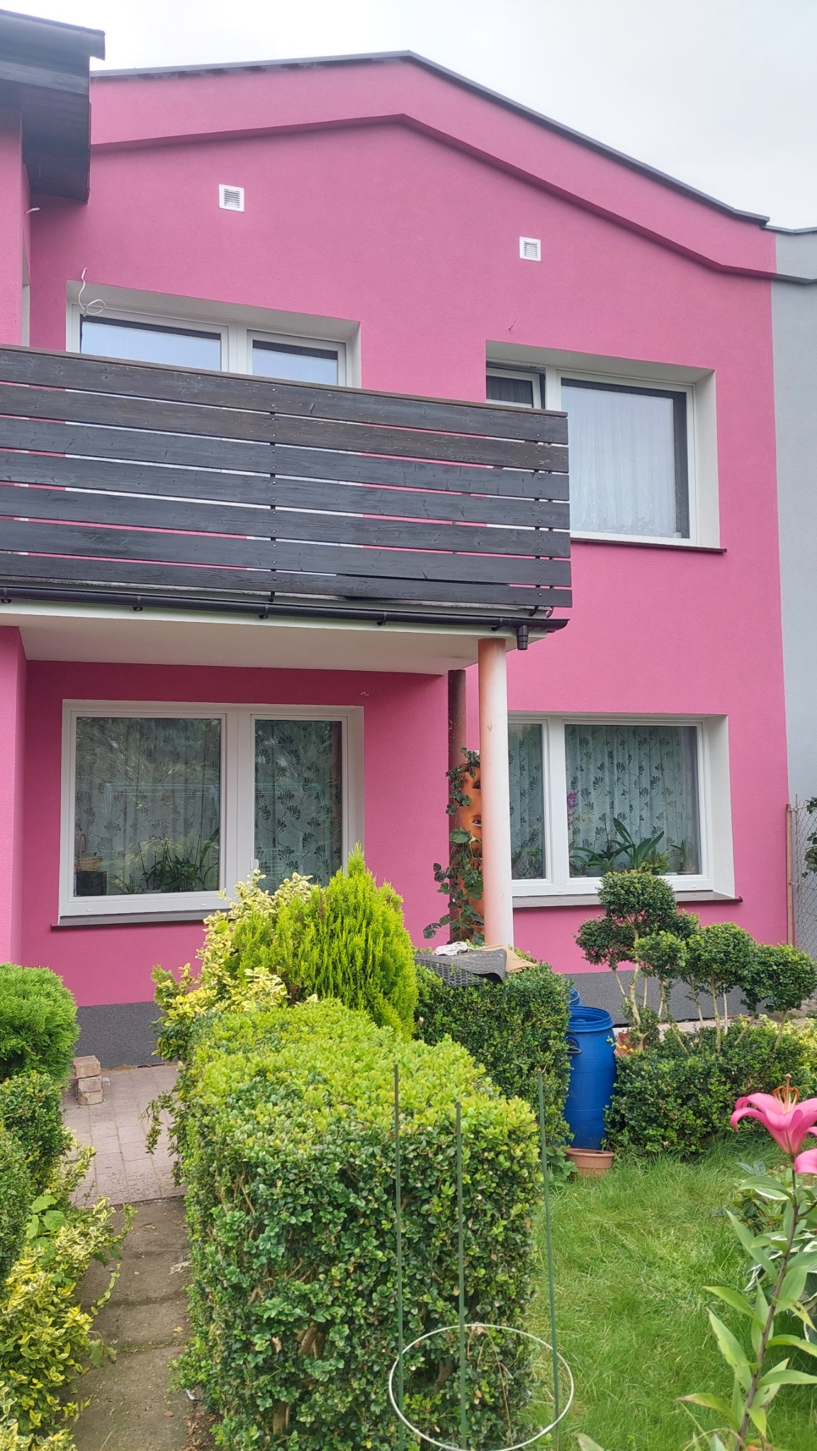 Dwupiętrowy dom z różową elewacją i ciemnym drewnianym balkonem, otoczony zielenią. Widoczne okna z firankami i niebieska beczka na deszczówkę w ogrodzie.