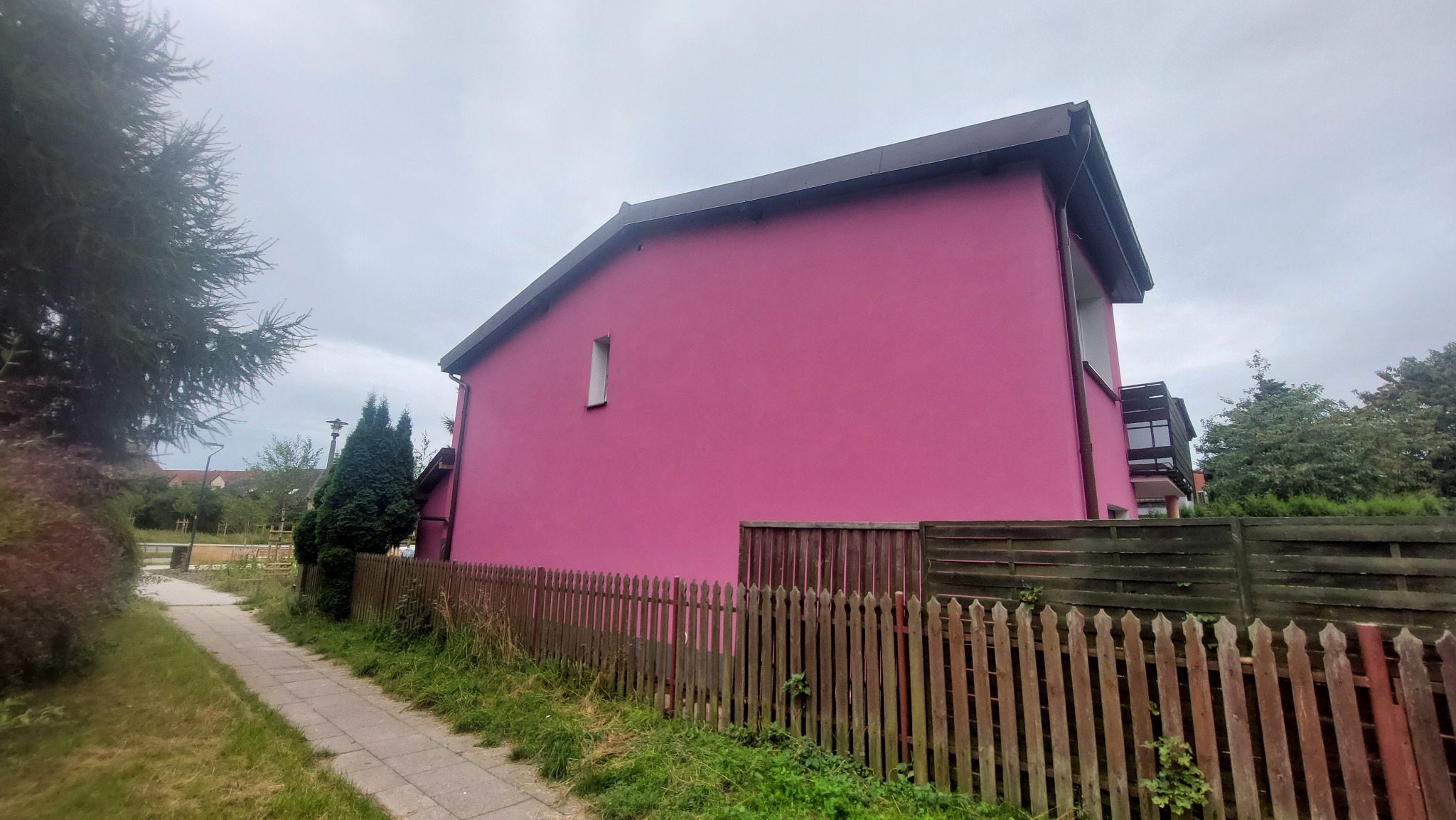Różowa elewacja dwukondygnacyjnego domu jednorodzinnego widziana z boku, z drewnianym płotem w pierwszym planie i zielenią w tle, pod pochmurnym niebem.