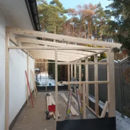 Konstrukcja szkieletowa drewnianej altany przylegającej do budynku, widoczne narzędzia, drabina i deski, częściowo ośnieżony teren.