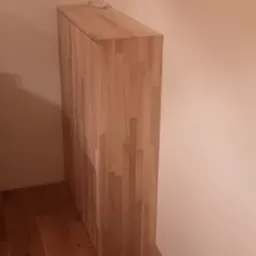 Narożna szafka z jasnego drewna klejonego, umieszczona w rogu pokoju z drewnianą podłogą, widoczne narzędzia i dywanik na podłodze.