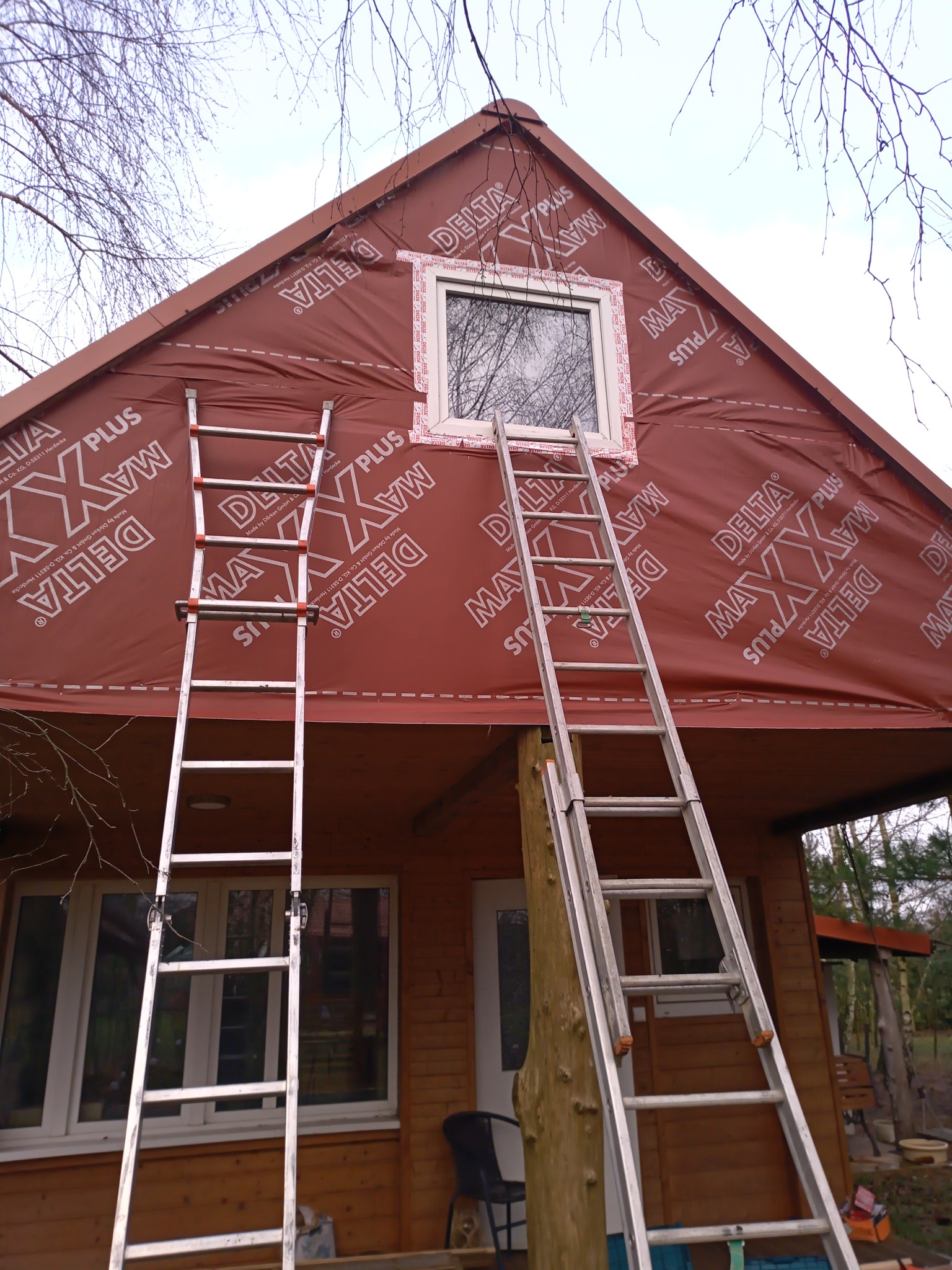 Dom z drewnianą elewacją w trakcie ocieplania, z widoczną membraną ochronną Delta Maxx Plus, nowo zamontowanym oknem i dwiema przystawionymi drabinami.