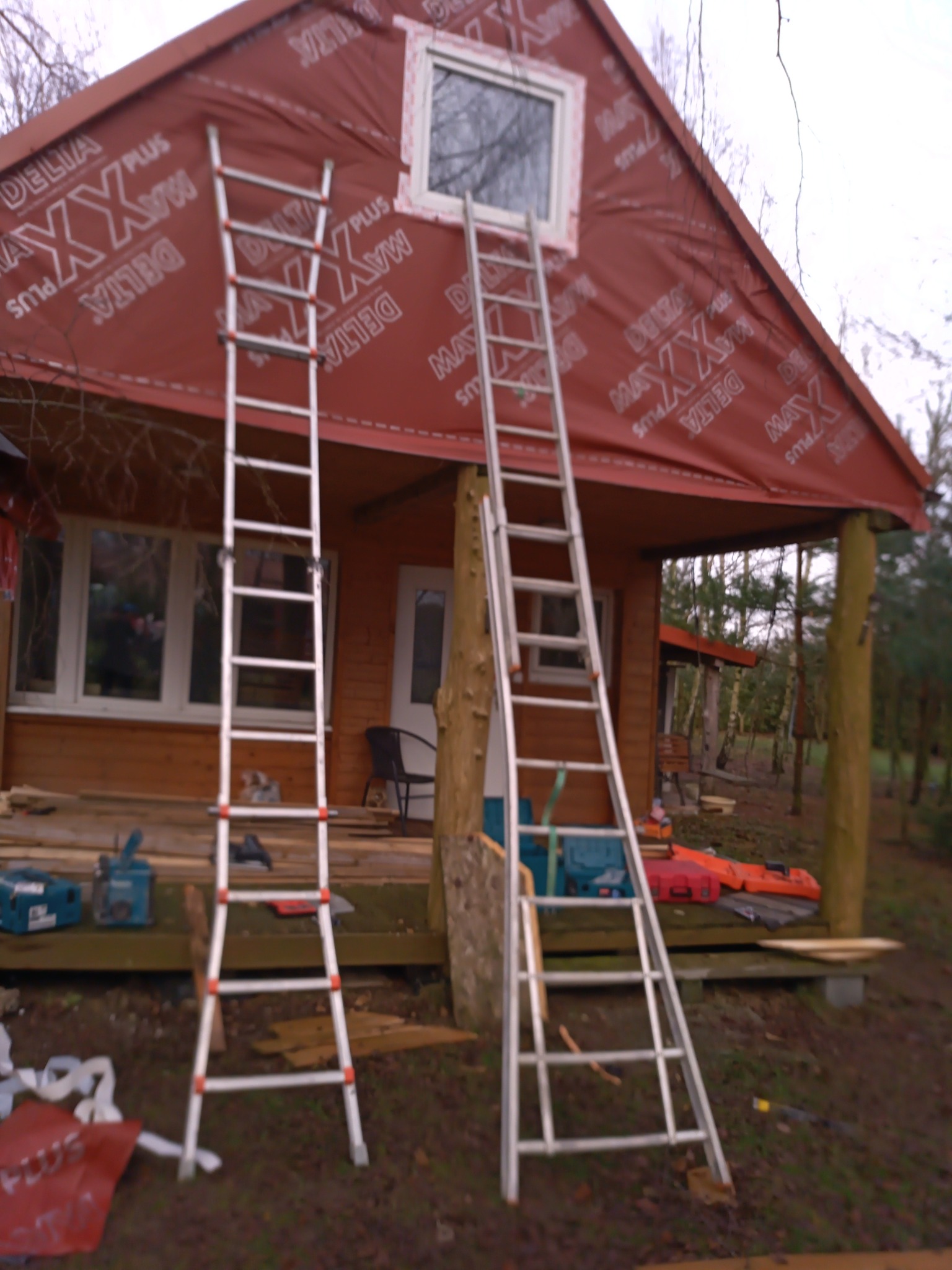 Drewniany domek z dwoma aluminiowymi drabinami opartymi o ścianę z oknem, pokrytą czerwoną folią z nadrukiem, widoczne narzędzia i skrzynki narzędziowe.