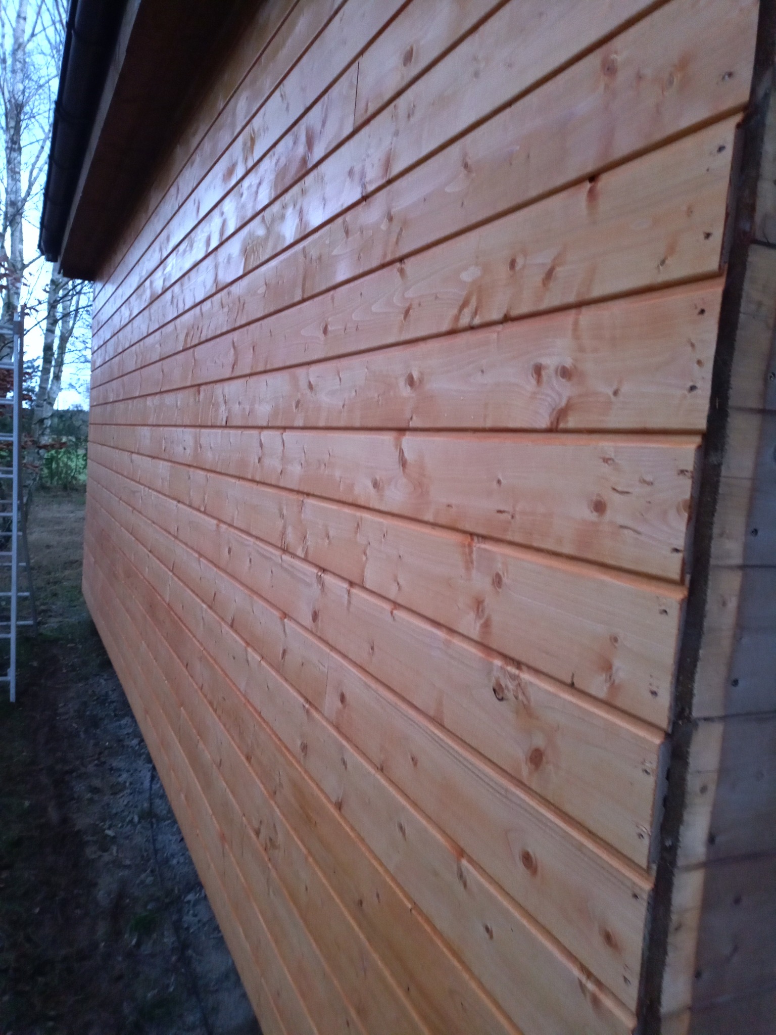 Świeżo odeskowana ściana budynku z jasnego drewna, widoczne sęki i usłojenie, zadaszenie i fragment drabiny w tle.