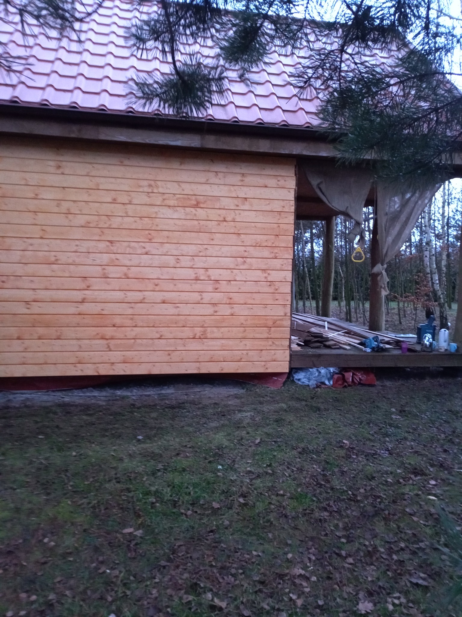 Fragment drewnianej konstrukcji z czerwoną dachówką, prawdopodobnie w trakcie budowy, z widocznymi narzędziami i materiałami w tle.