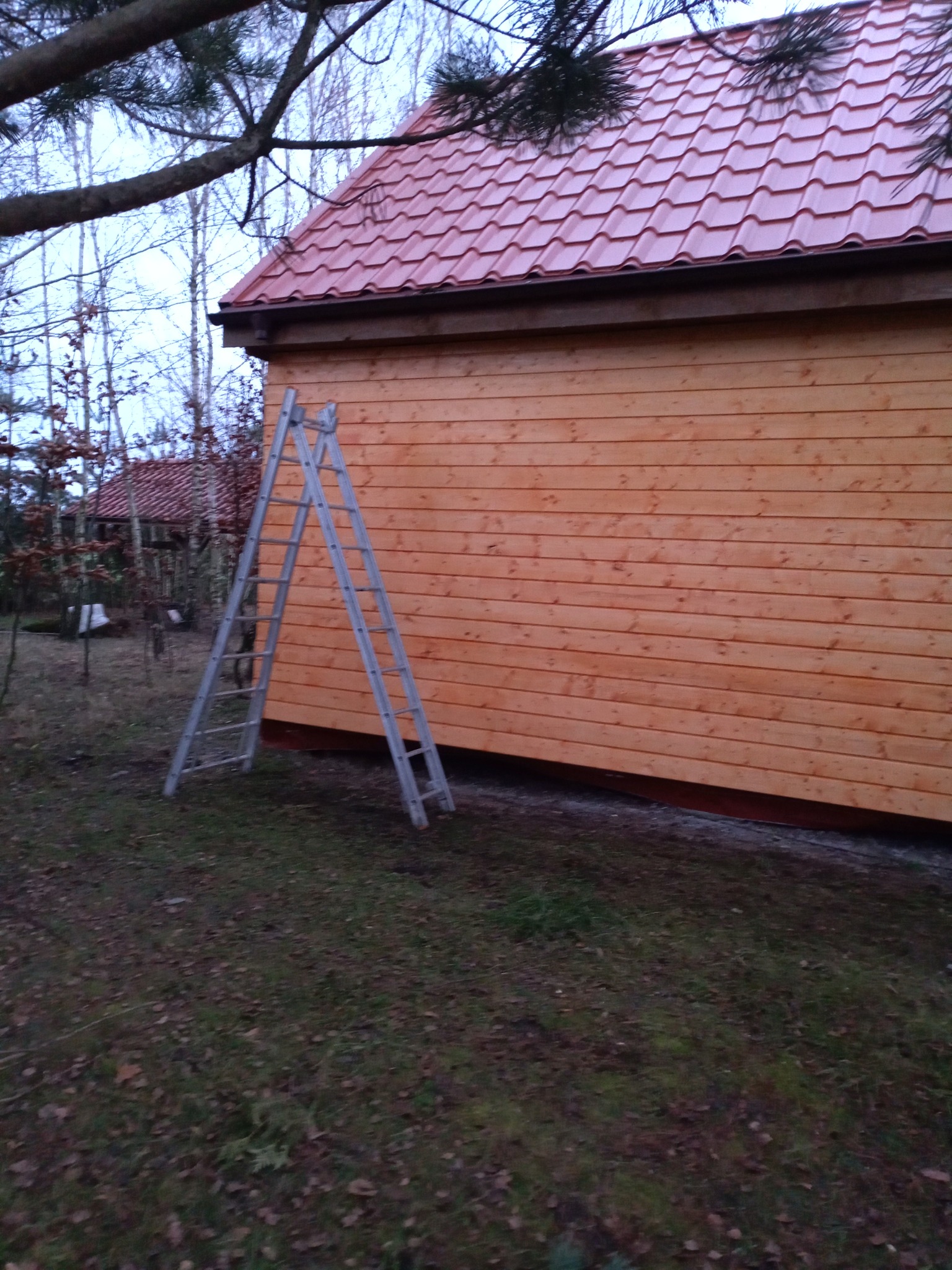 Nowa drewniana altana z czerwoną dachówką, widoczna drabina oparta o ścianę, w tle drzewa bez liści.