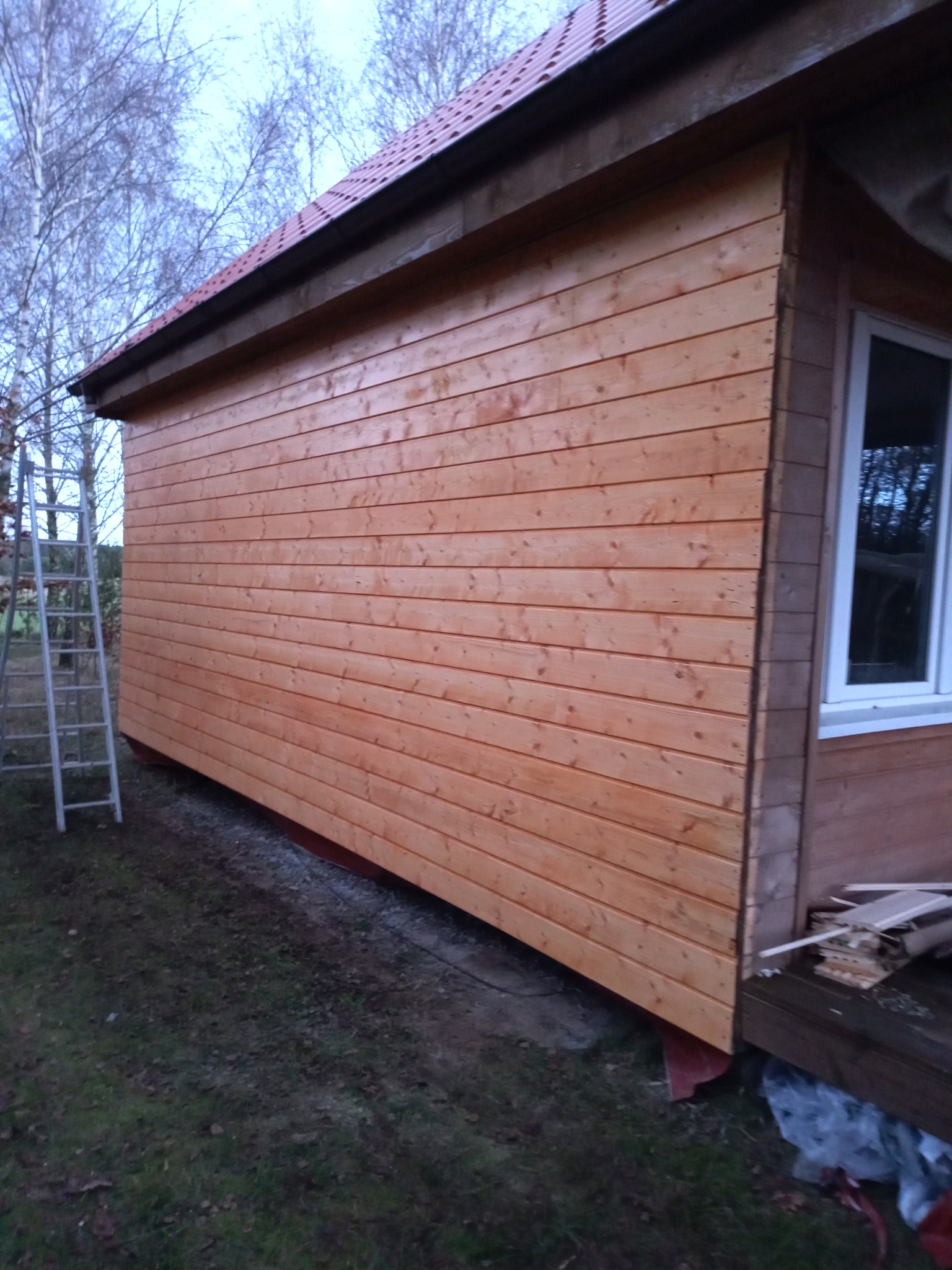 Świeżo wybudowana altana z jasnego drewna, widoczna drabina oparta o ścianę, podłoże częściowo wyłożone kamieniami.