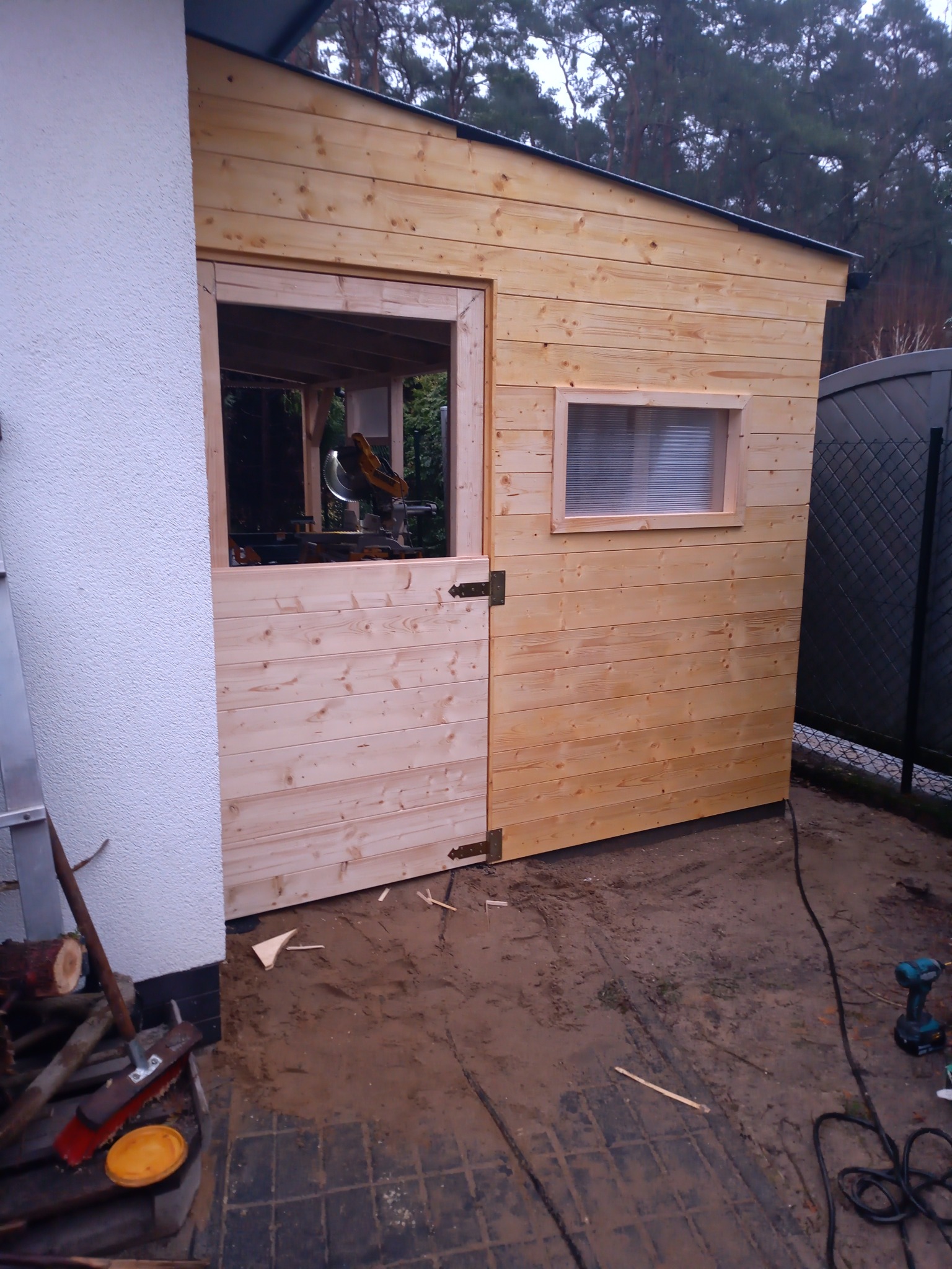 Surowa drewniana konstrukcja altany w trakcie budowy, widoczna piła stołowa wewnątrz, ziemiste podłoże, narzędzia i drewno w pobliżu ściany budynku.