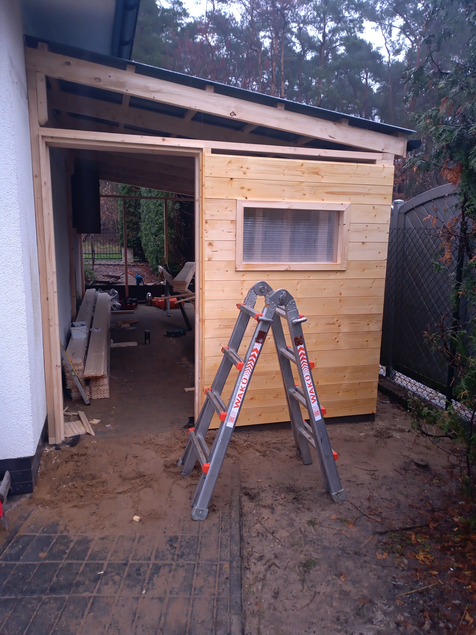 Drewniana konstrukcja altany w trakcie budowy, widoczna drabina aluminiowa, deski i narzędzia wewnątrz. Surowy stan wykończenia, widoczne elementy konstrukcyjne i okno z przezroczystą folią.