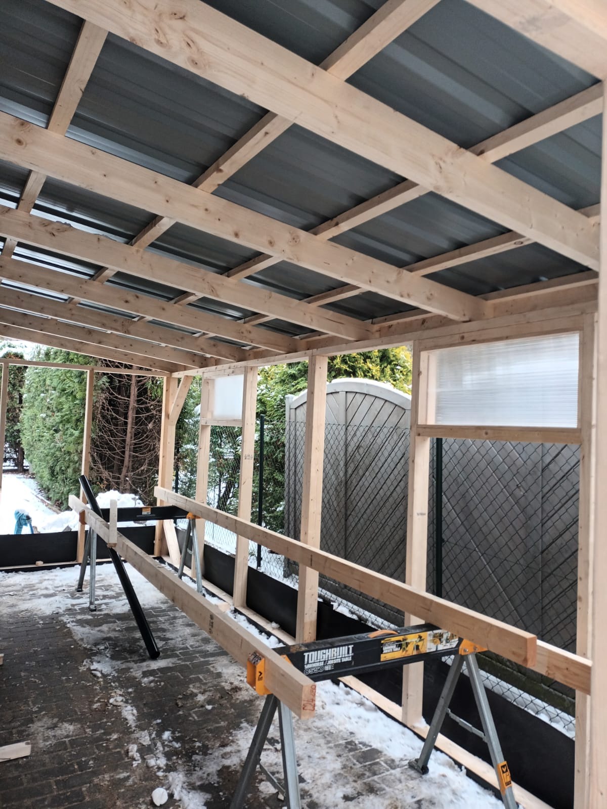 Konstrukcja z surowego drewna z metalowym dachem w trakcie budowy, prawdopodobnie zadaszenie tarasu lub weranda, widoczne narzędzia i śnieg na ziemi.