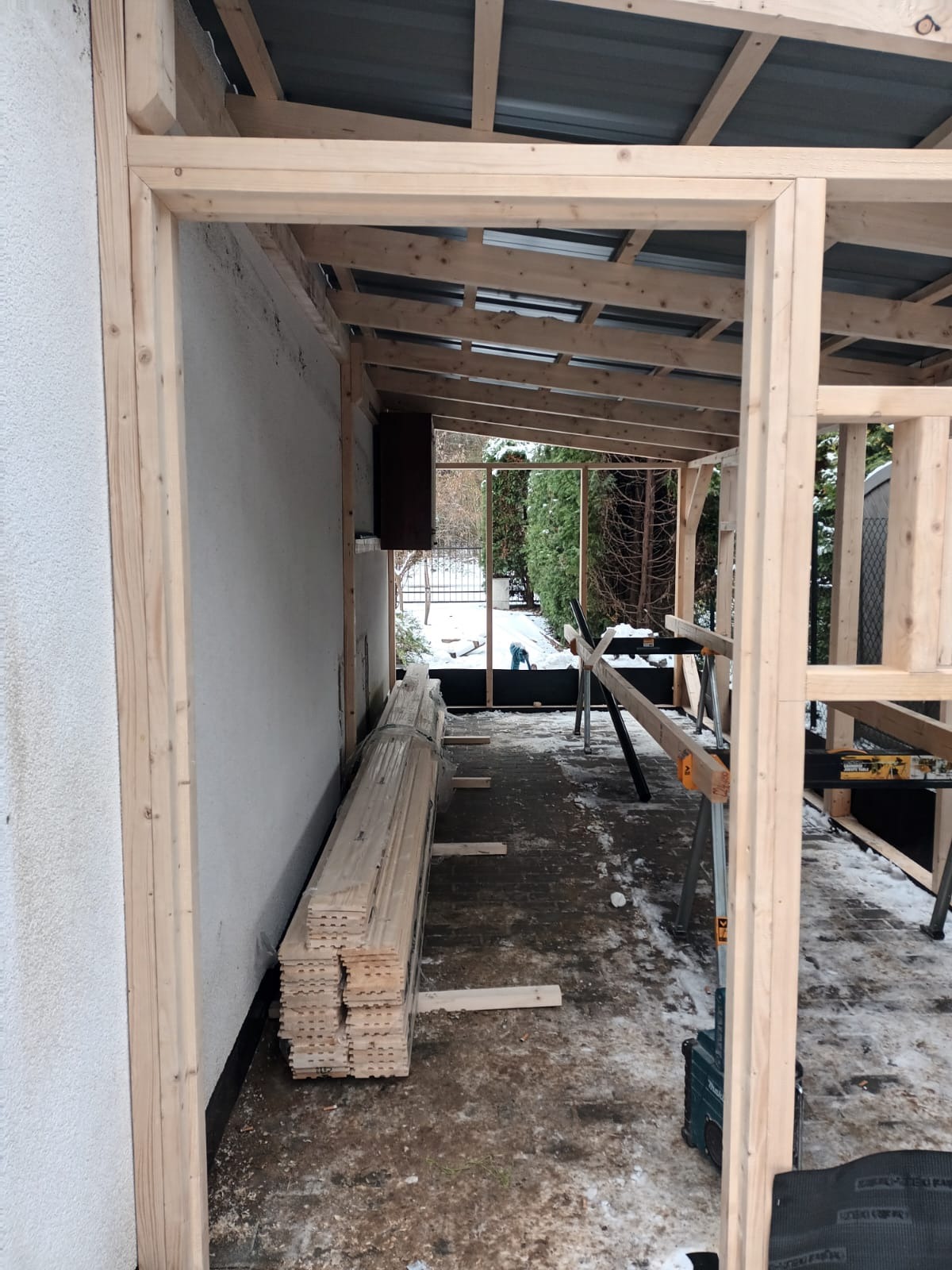 W trakcie budowy drewnianej konstrukcji z widocznym stosem desek, elementami szkieletu i zadaszeniem, częściowo na zewnątrz budynku w zimowej scenerii.