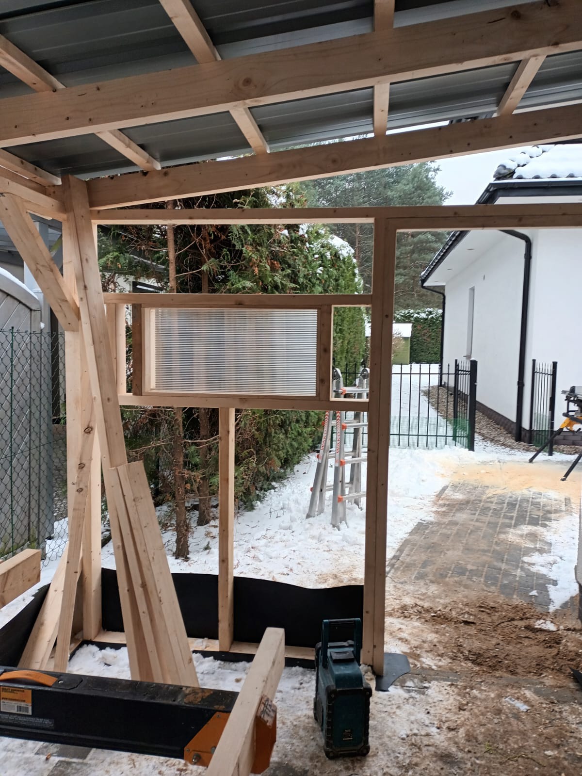 Wznoszenie drewnianej konstrukcji altany z częściowo zamontowaną przezroczystą ścianą, widoczne narzędzia i resztki materiałów budowlanych na ziemi pokrytej śniegiem.