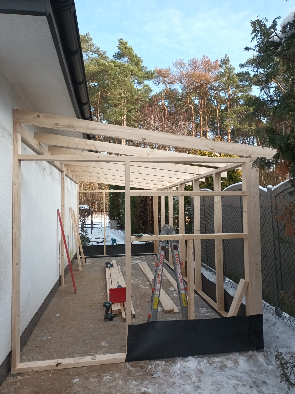 Konstrukcja szkieletowa drewnianej altany przylegającej do budynku, widoczne narzędzia, drabina i deski, częściowo ośnieżony teren.