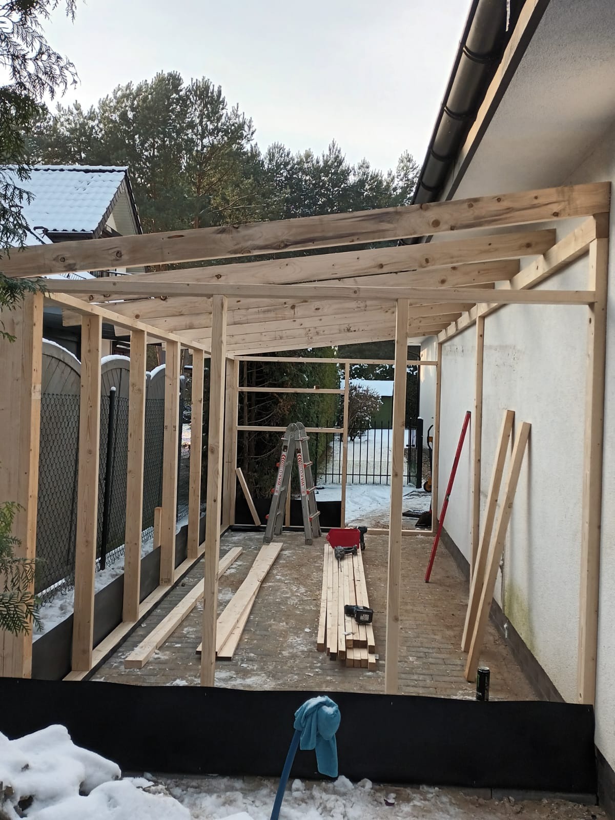 Konstrukcja drewniana altany w trakcie budowy, widoczne deski, drabina, poziomica i narzędzia na tle zimowego krajobrazu.