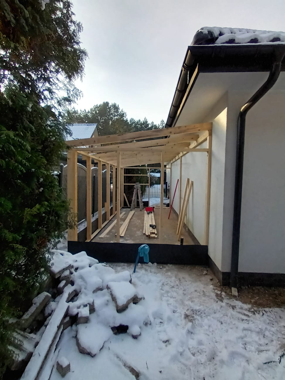 Konstrukcja drewniana w trakcie budowy, prawdopodobnie zadaszenia tarasu lub wiaty, widoczne elementy szkieletu i przygotowane deski, zimowa sceneria z śniegiem na ziemi i gruzie.