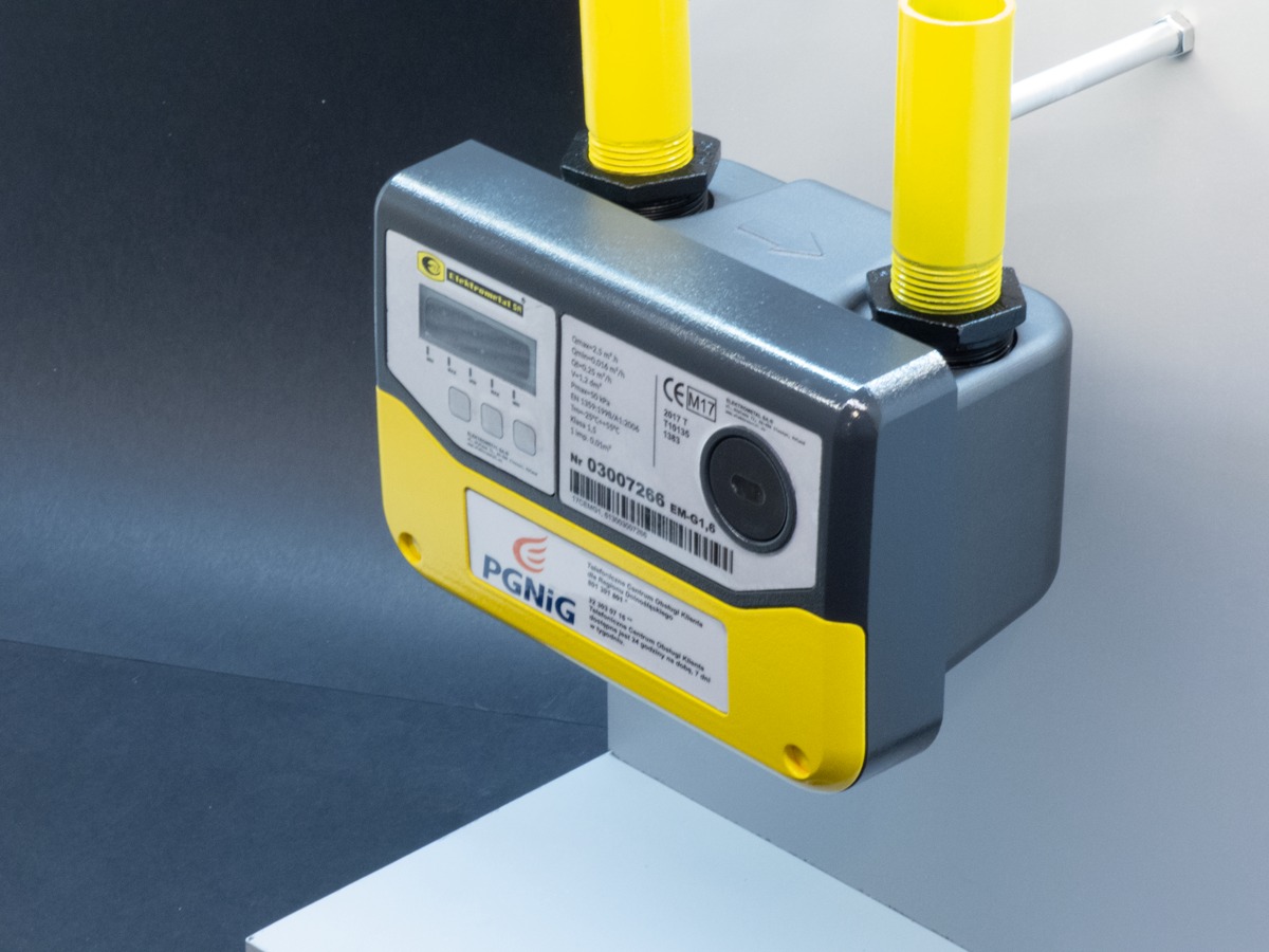 Szary gazomierz z żółtymi elementami i rurami, zamontowany na ścianie, z widocznymi parametrami technicznymi i logo PGNIG.