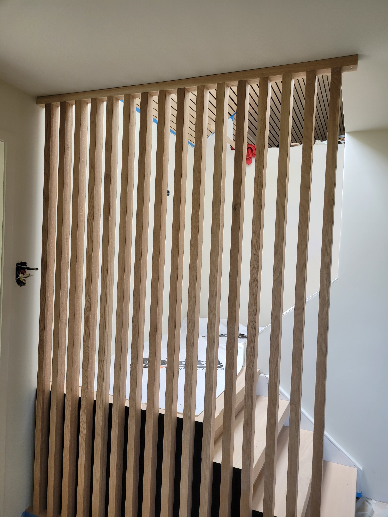 Drewniana ażurowa ścianka działowa z pionowych listew oddzielająca schody od korytarza, widoczne niedokończone prace wykończeniowe i brakująca osłona gniazdka elektrycznego.