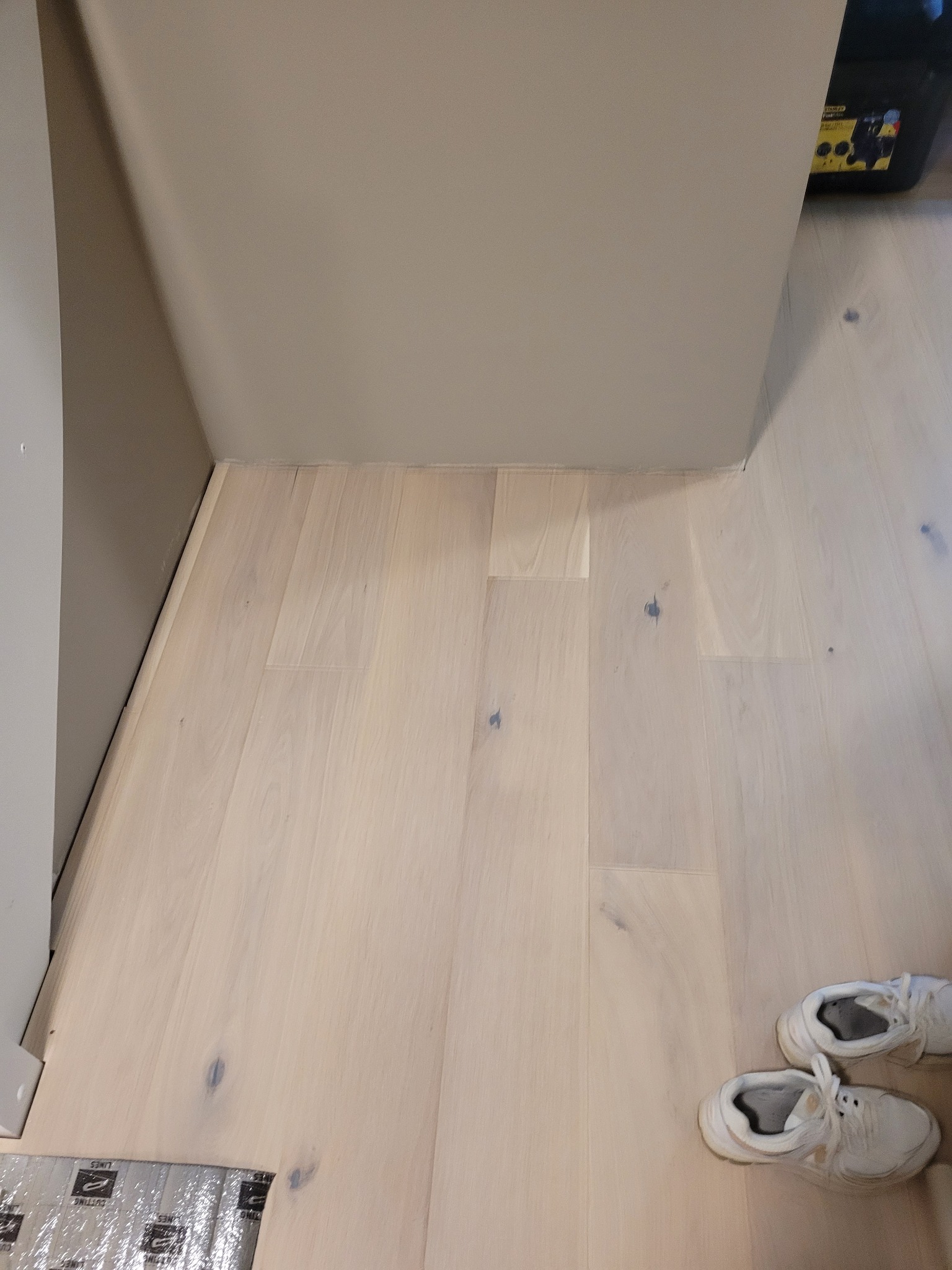 Jasne, drewniane panele podłogowe ułożone w kącie pomieszczenia, z widocznymi fragmentami folii ochronnej i parą białych butów w prawym dolnym rogu.